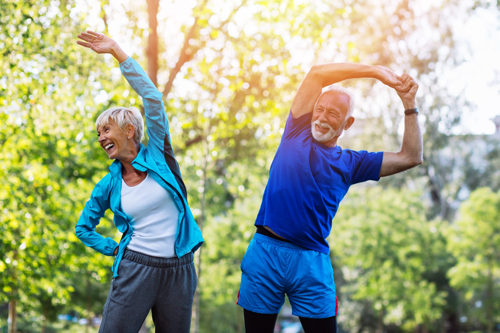 Two elderly individuals, one male and one female, are performing side stretches outdoors. They are smiling and dressed in athletic attire, with the woman in a light blue jacket and grey pants, and the man in a blue shirt and shorts. Lush greenery surrounds them.
