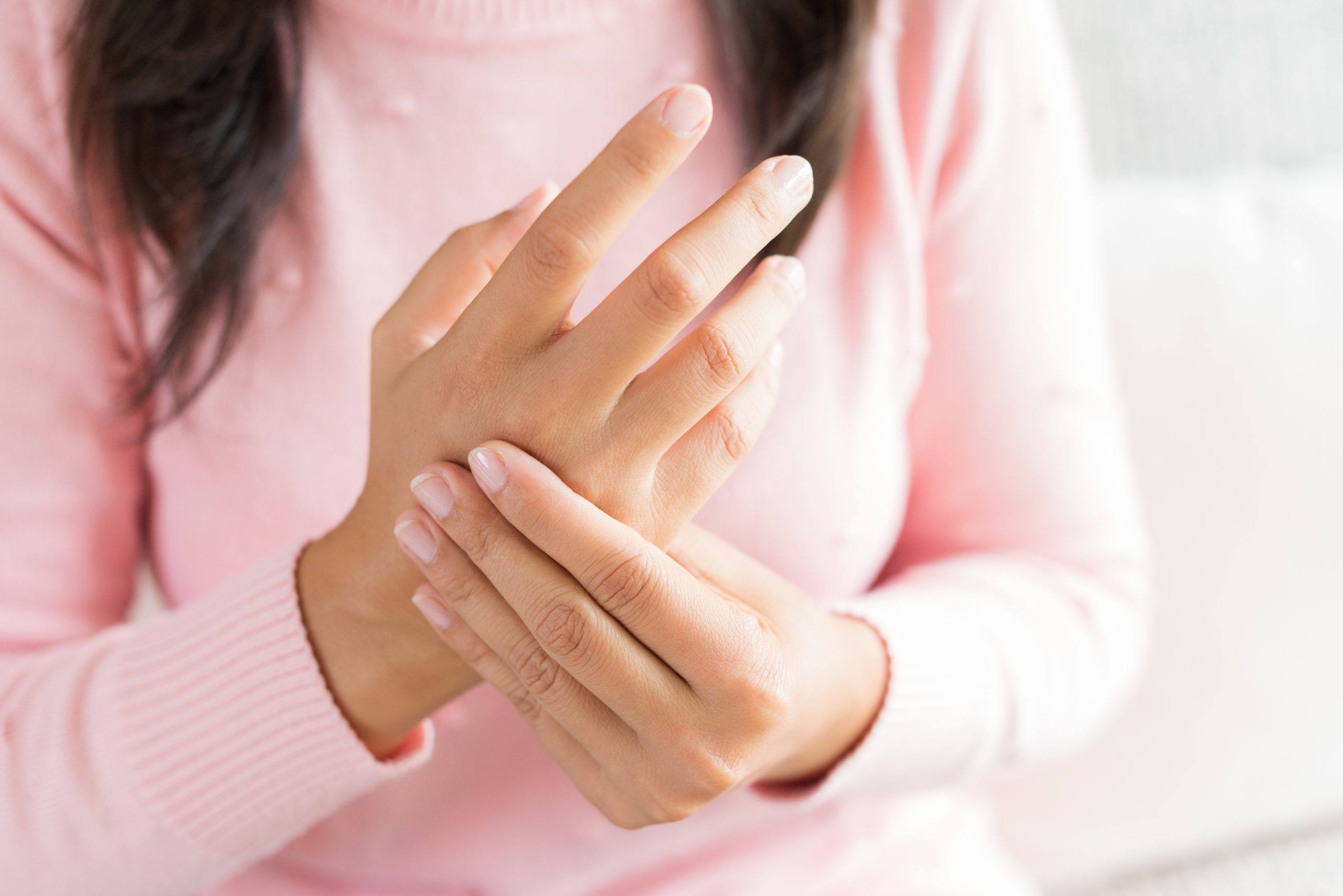 Close-up of a person wearing a pink long-sleeve top, holding their left hand with their right hand, appearing to massage the fingers. The background is blurred, focusing on the hands and partial upper torso.