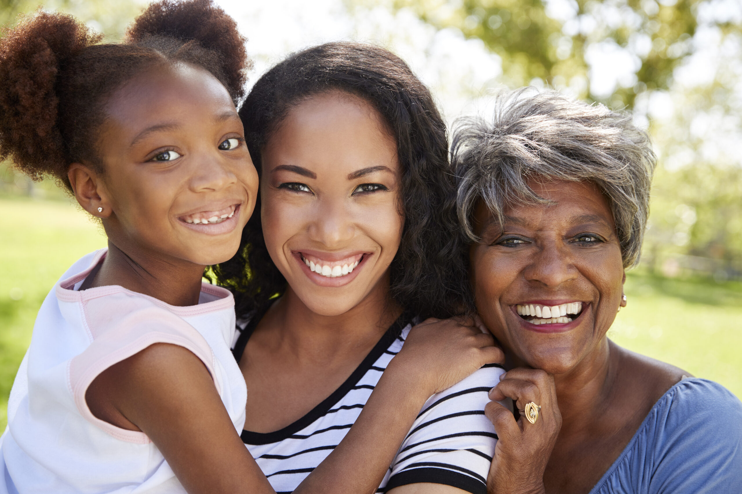 Three generations of women smiling and embracing outdoors. On the left is a young girl with puffy pigtails, in the center is a woman with long hair, and on the right is an older woman with short gray hair. Bright daylight and greenery provide a cheerful background.