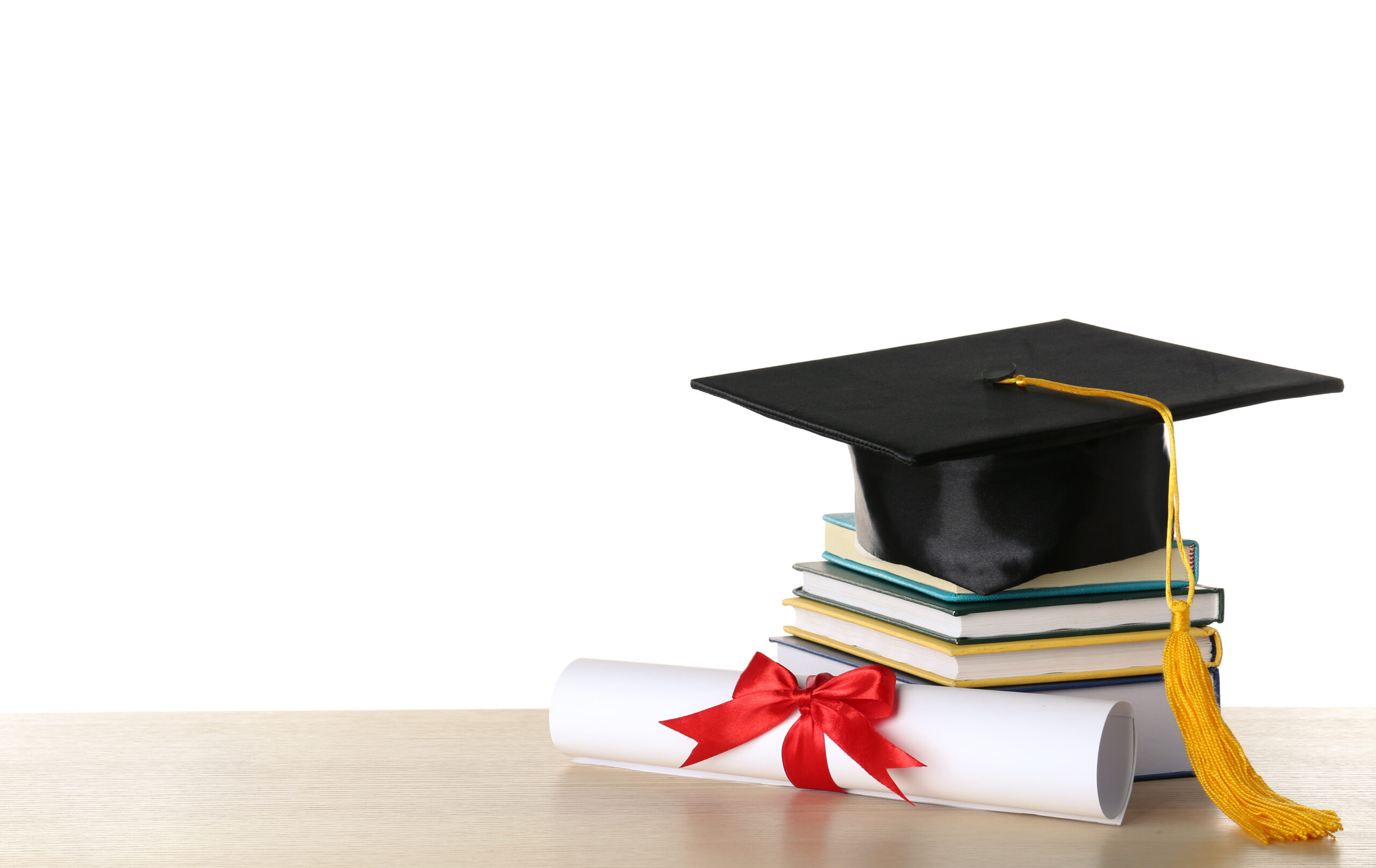 A graduation cap with a yellow tassel sits on top of a stack of books next to a rolled-up diploma tied with a red ribbon, resting on a wooden surface. The background is white, creating a clean and minimalistic look.