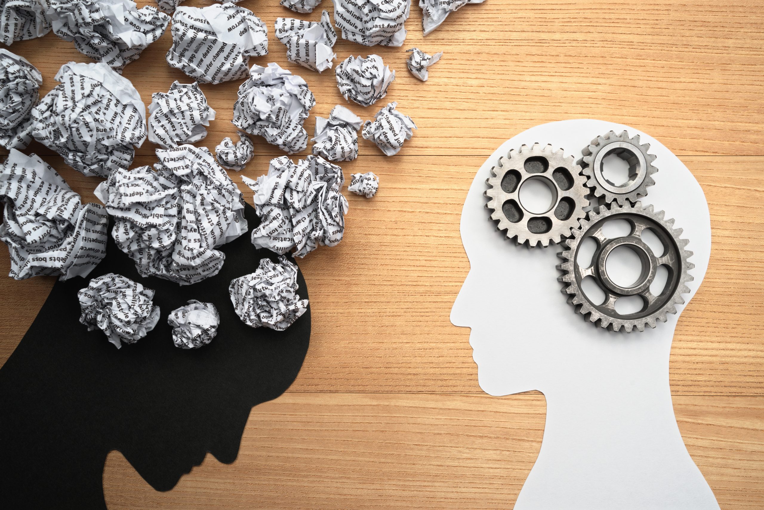 A conceptual image showing two paper cutout silhouettes of heads on a wooden surface. The left head is black with crumpled paper inside, transitioning to the right head, which is white with interlocking cogs inside, illustrating a transfer of ideas or thoughts.
