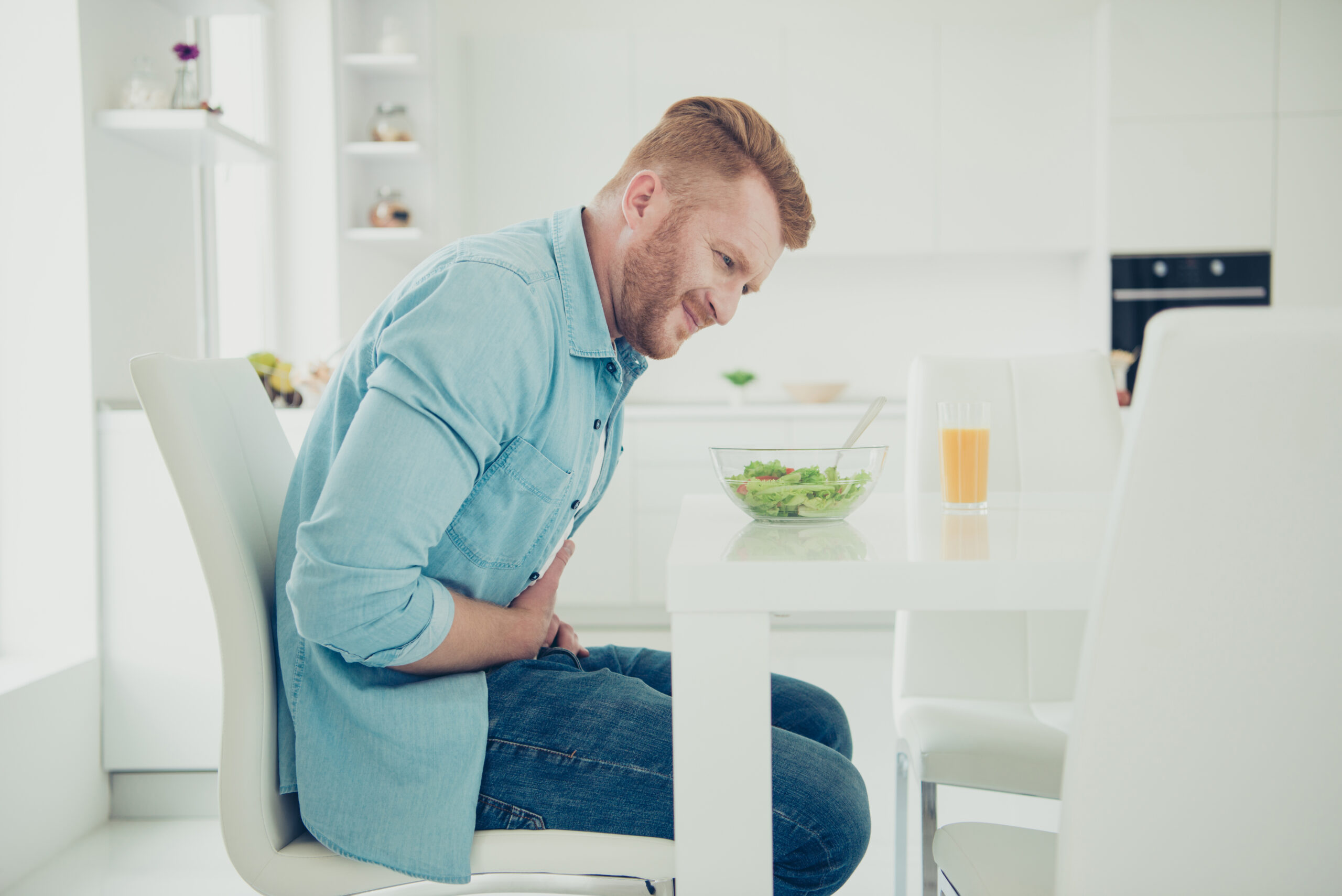 A man with red hair and a beard, wearing a light blue shirt and jeans, sits at a white kitchen table holding his stomach in discomfort. In front of him is a bowl of salad and a glass of orange juice. The kitchen is modern and predominantly white.