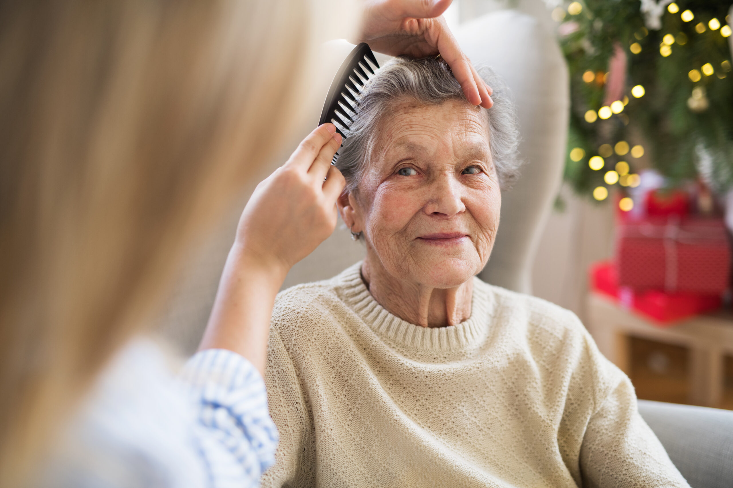 An elderly woman with gray hair and wearing a beige sweater sits in a chair, smiling contentedly as another person combs her hair. A decorated Christmas tree and wrapped gifts are visible in the background, indicating a festive setting.