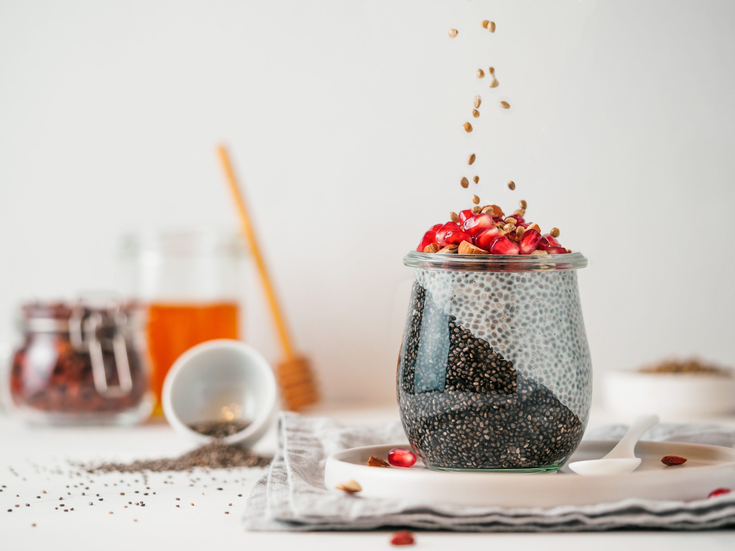 A glass jar filled with chia pudding is topped with pomegranate seeds and nuts, with some of the toppings being sprinkled over it. The jar is placed on a white plate with a spoon next to it. In the background, there are jars of honey and seeds, and a blurred wooden spoon.