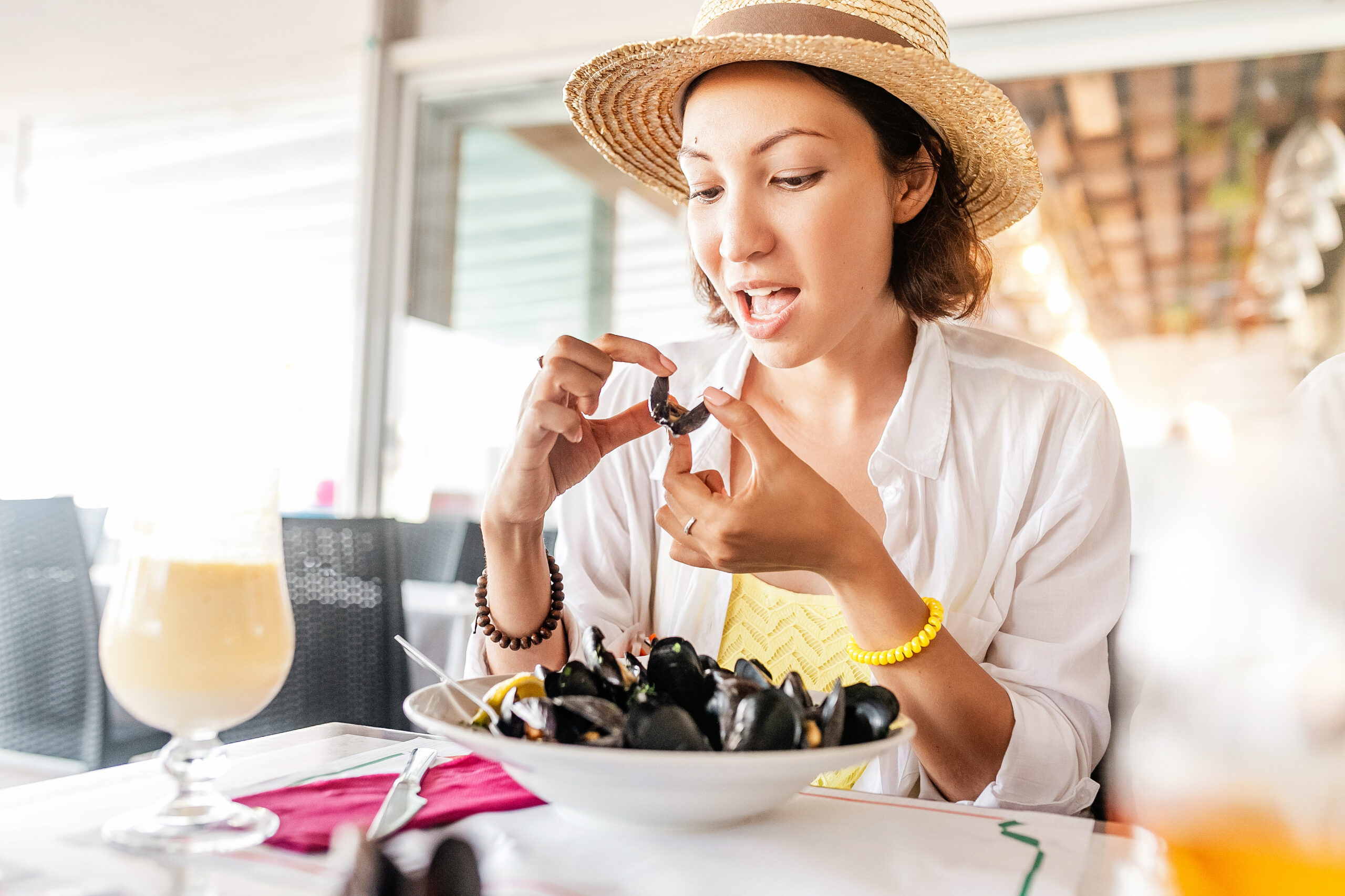 A woman wearing a straw hat and white shirt is seated at a table in a restaurant, eating mussels. In front of her is a bowl filled with mussels, and a drink is on the table. She appears to be enjoying her meal, holding a mussel shell in her hands.