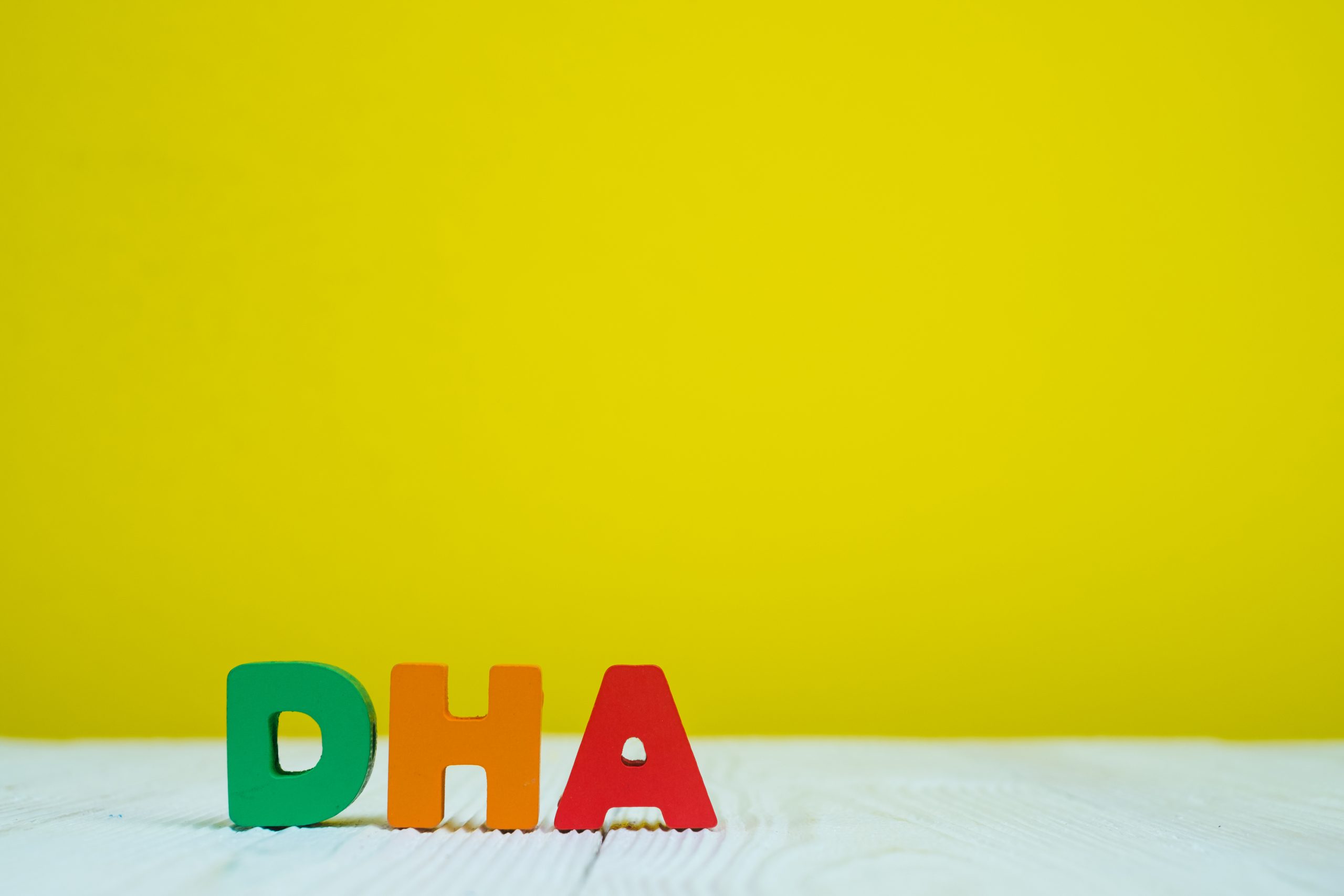 Colorful wooden letters spelling "DHA" are arranged on a light wooden surface against a vibrant yellow background. The letters are painted green, orange, and red, respectively, and are positioned in a neat row from left to right.