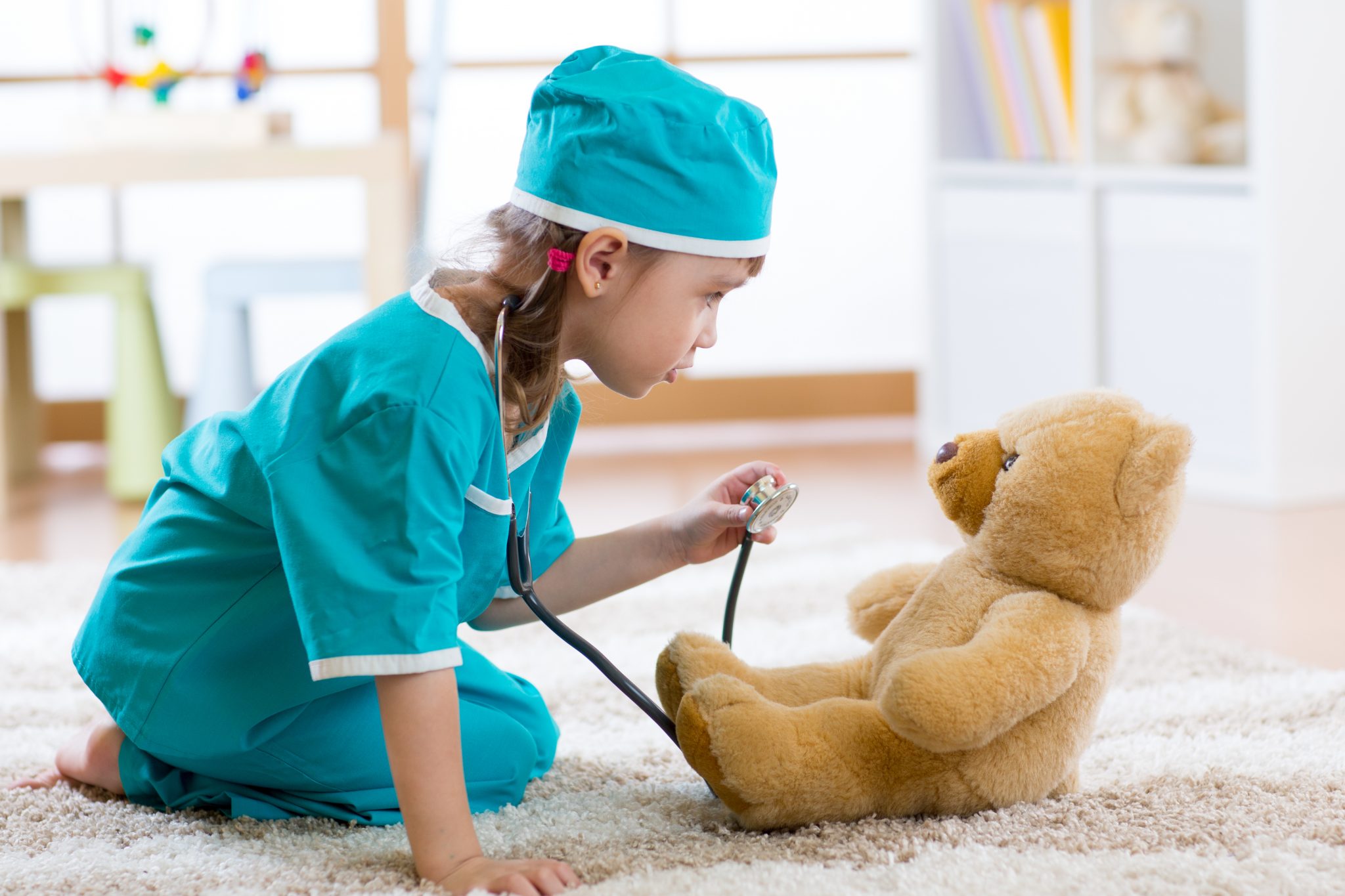 A young child dressed in teal medical scrubs and a matching cap kneels on a carpeted floor using a stethoscope to check a teddy bear. The room has a bright, playful atmosphere with toys in the background.