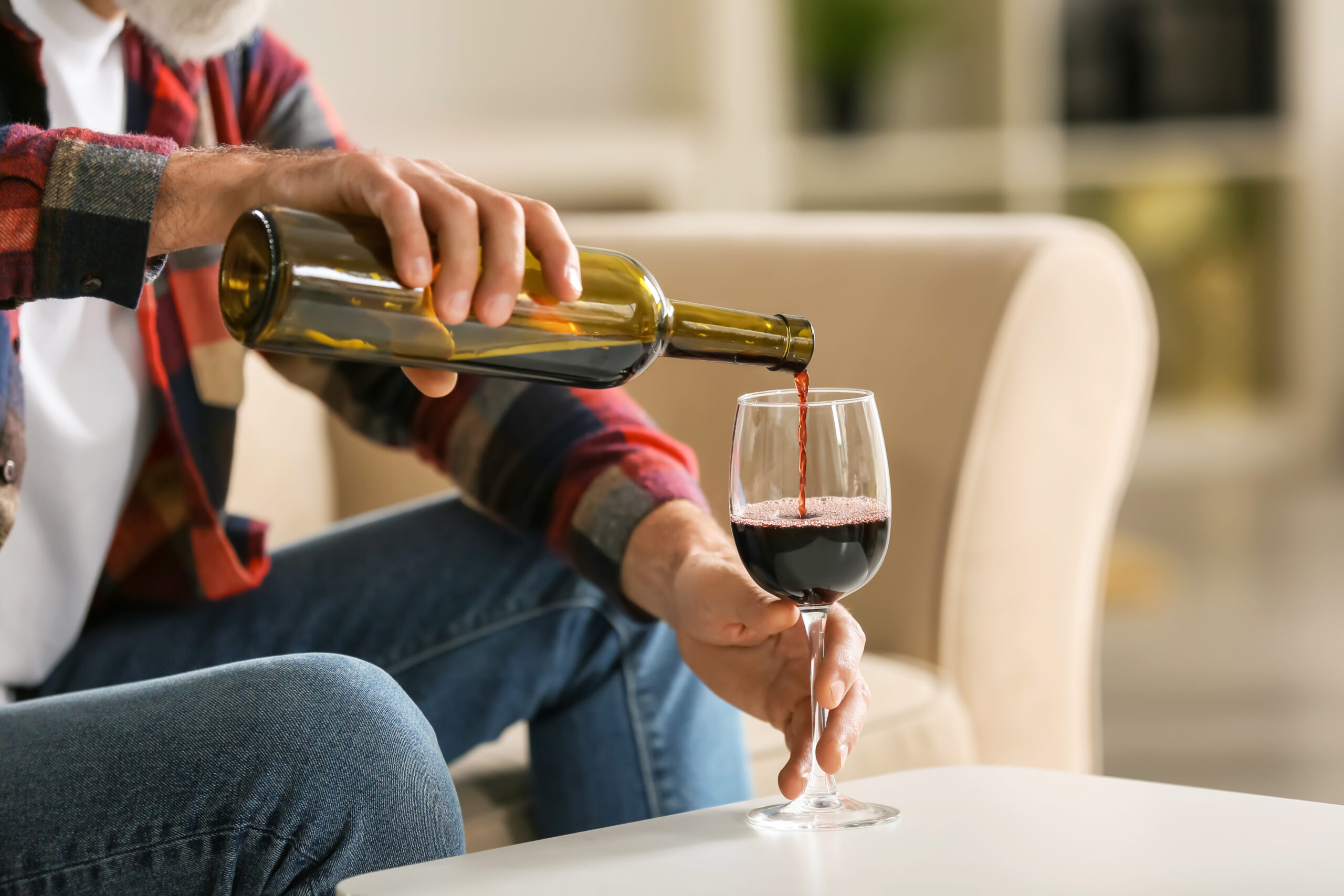 A person is sitting on a couch, wearing a red plaid shirt and blue jeans, and pouring red wine from a bottle into a wine glass. The scene takes place in a cozy living room with neutral-colored furniture in the background.