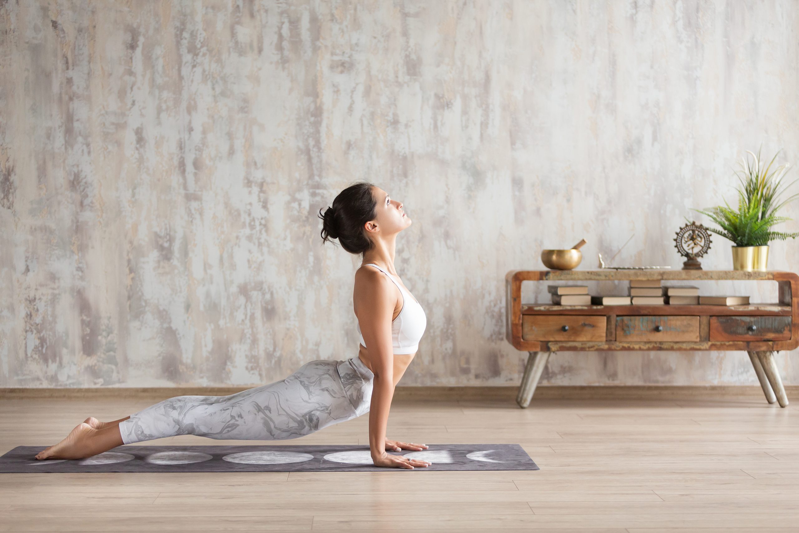 A woman practices yoga in a spacious, minimalist room with wooden flooring. She is performing the upward-facing dog pose on a yoga mat. Light enters through a window out of frame, illuminating the scene. A wooden side table with decorative items is in the background.