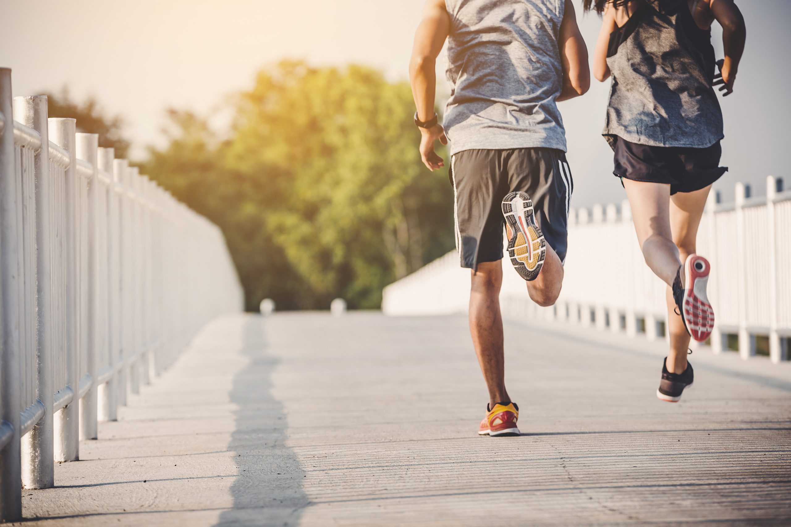 Two people are jogging on a paved path with white railings on both sides. They are wearing athletic clothing and sneakers and running away from the camera. The background shows greenery and a slightly hazy sky, suggesting early morning or late afternoon light.