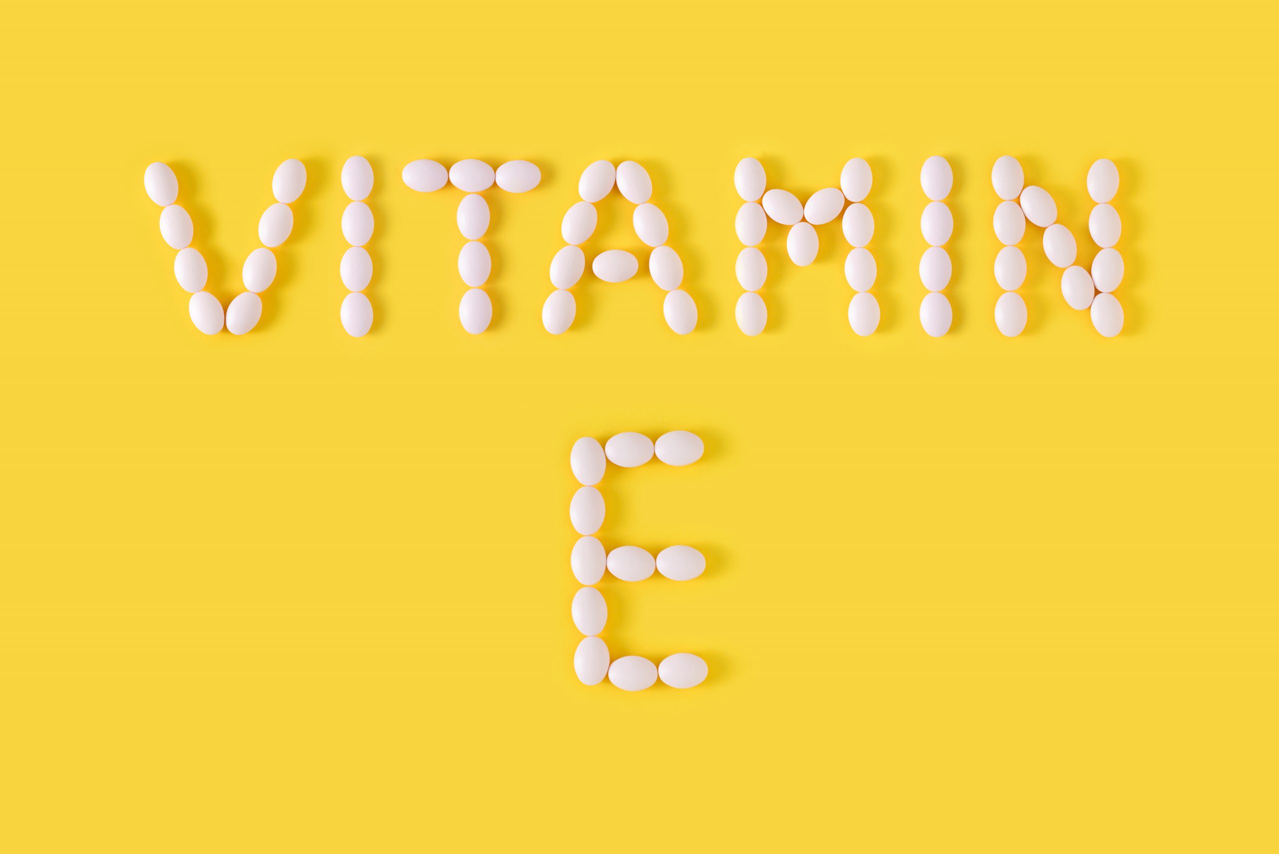 The text "VITAMIN E" is spelled out using white pills on a bright yellow background.