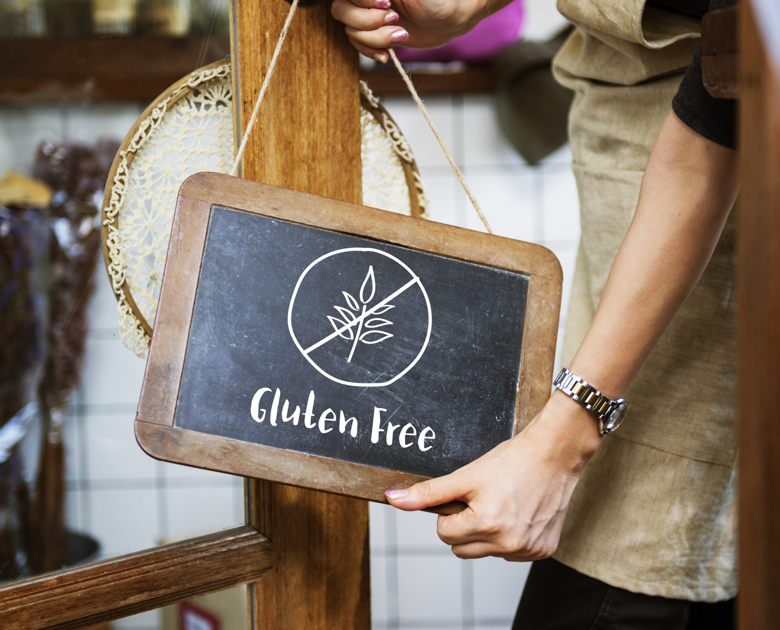 A person wearing an apron is holding a small chalkboard sign hanging on a door. The sign has a symbol of a crossed-out wheat stalk and reads "Gluten Free" underneath, indicating that the establishment or items offered are gluten-free.
