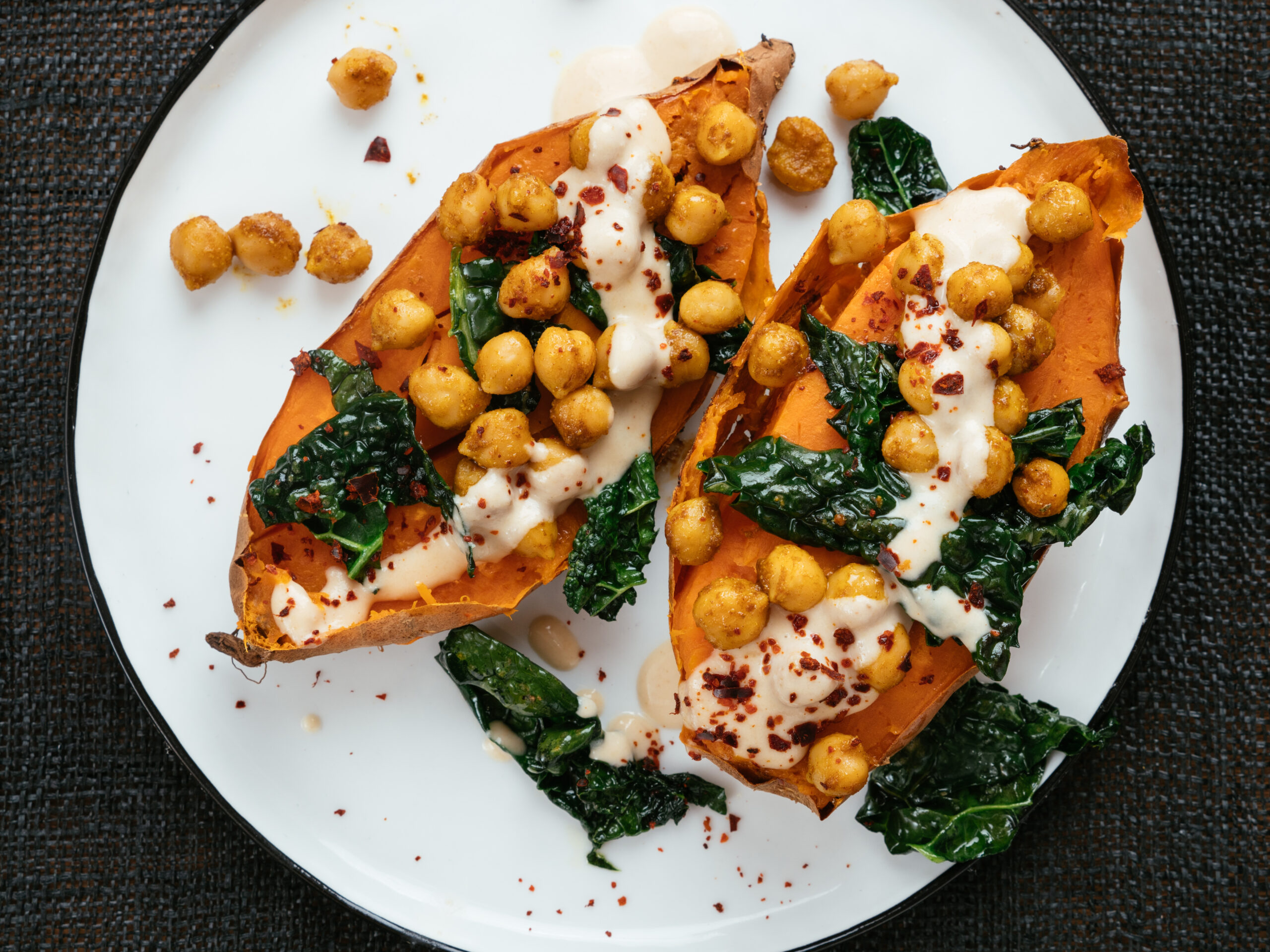 A white plate featuring roasted sweet potato halves topped with crispy chickpeas, sautéed kale, drizzled with a creamy sauce, and sprinkled with red pepper flakes. The dish is arranged on a dark, textured surface.