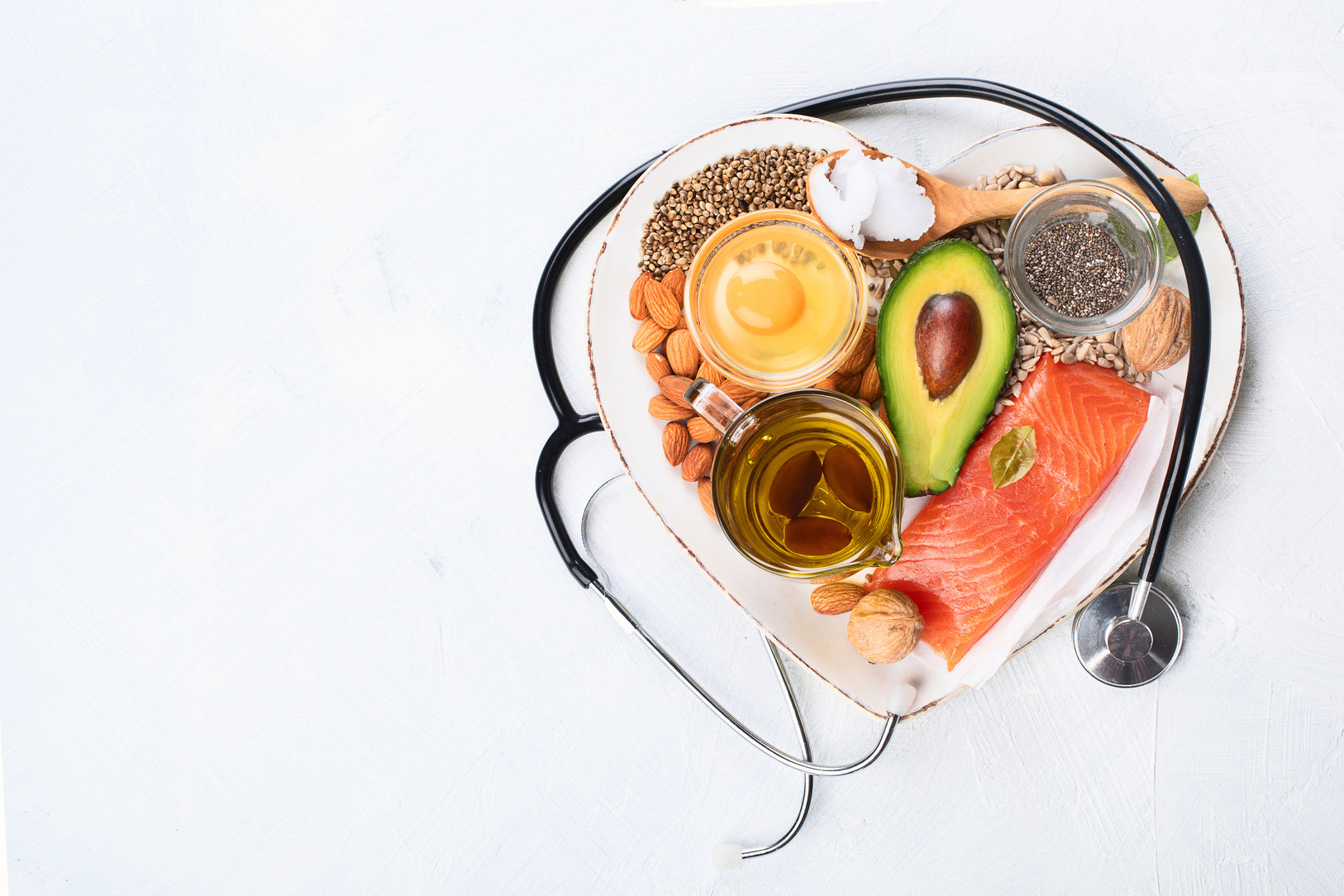 A heart-shaped dish containing various healthy foods including salmon, avocado, almonds, chia seeds, walnuts, olive oil, and an egg yolk. A stethoscope is placed around the dish, symbolizing the importance of heart health.