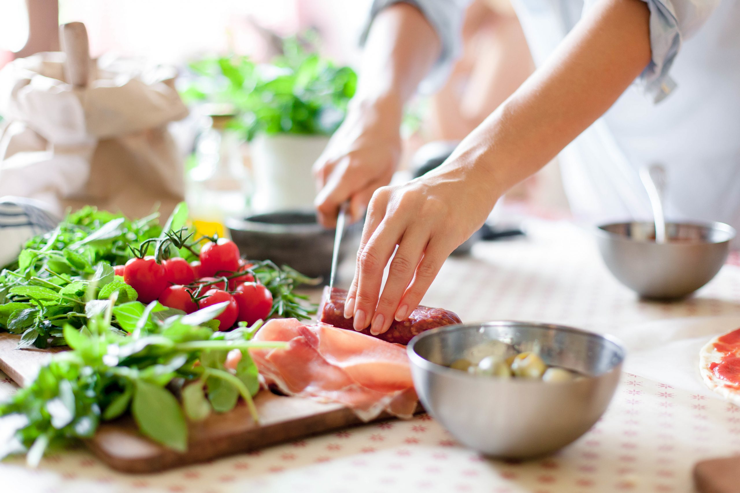 A person slicing meat on a cutting board surrounded by various fresh ingredients, including cherry tomatoes, herbs, and leafy greens. Bowls and a paper bag are on the table, suggesting food preparation in a bright kitchen.