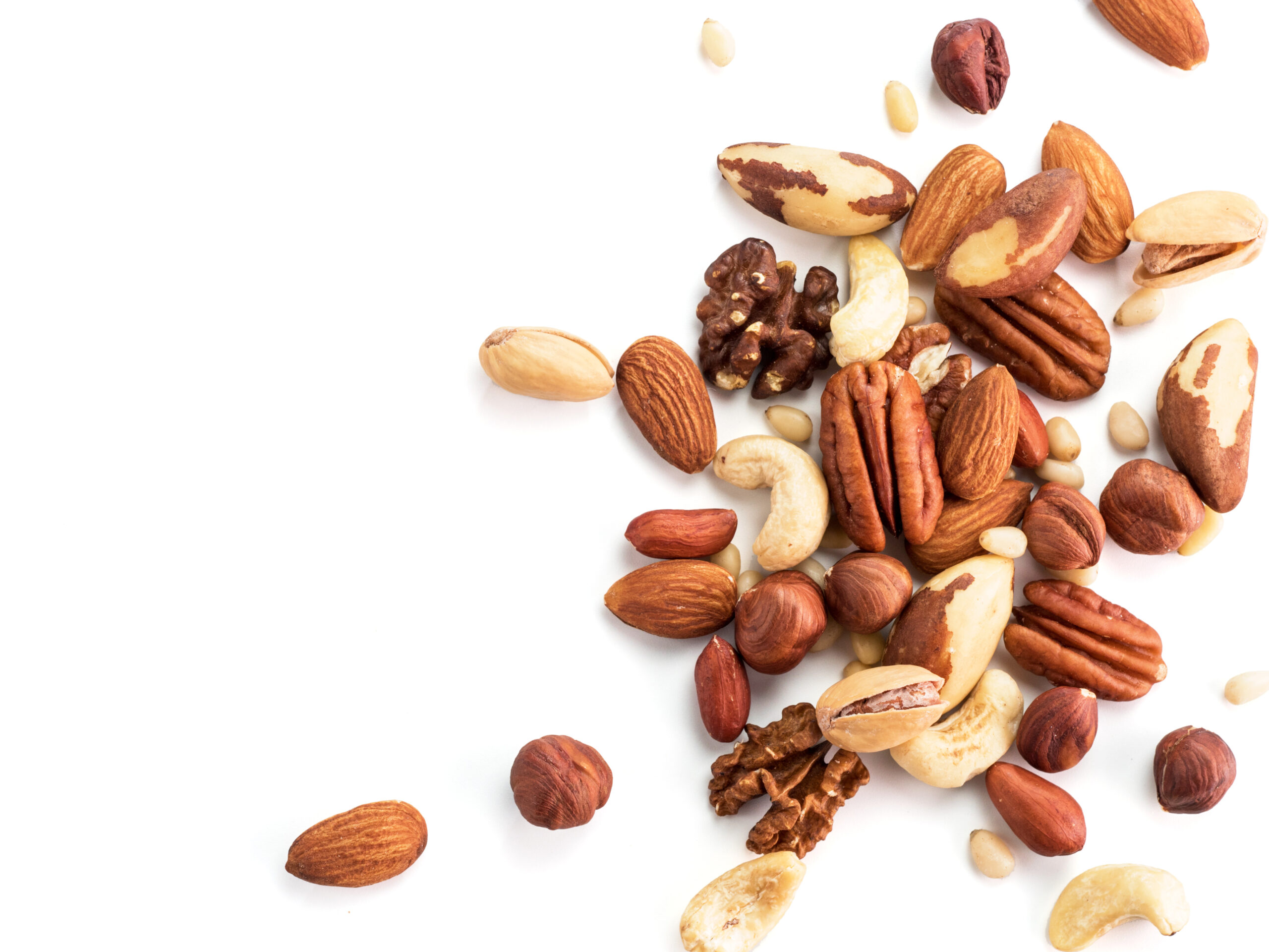 A scattered pile of mixed nuts including almonds, cashews, walnuts, pecans, hazelnuts, and Brazil nuts against a white background. Some nuts are whole, while others are shelled.