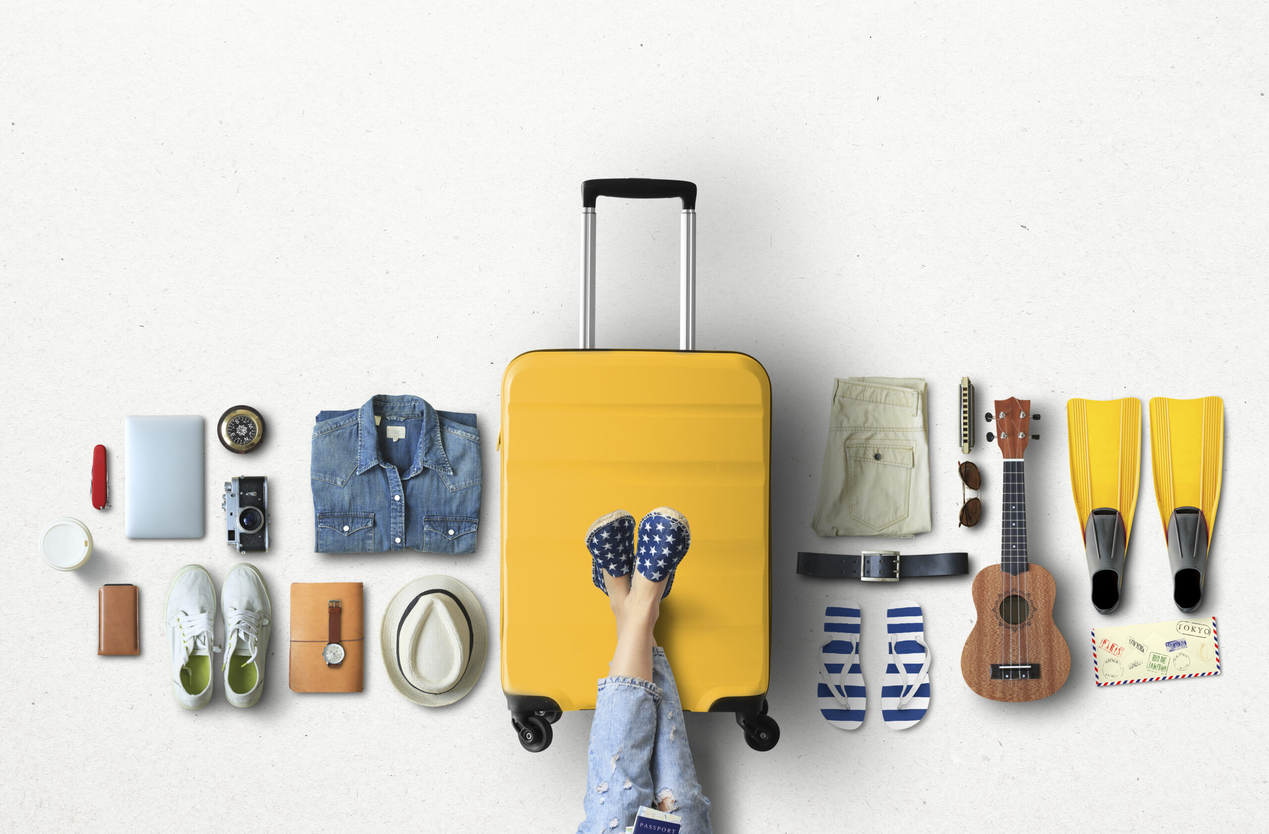 Overhead view of various travel items neatly arranged around a central yellow suitcase. Included are a camera, sneakers, hat, passport, notebook, denim shirt, rolled pants, watch, ukulele, snorkeling fins, and a woman's crossed legs in polka dot shoes.