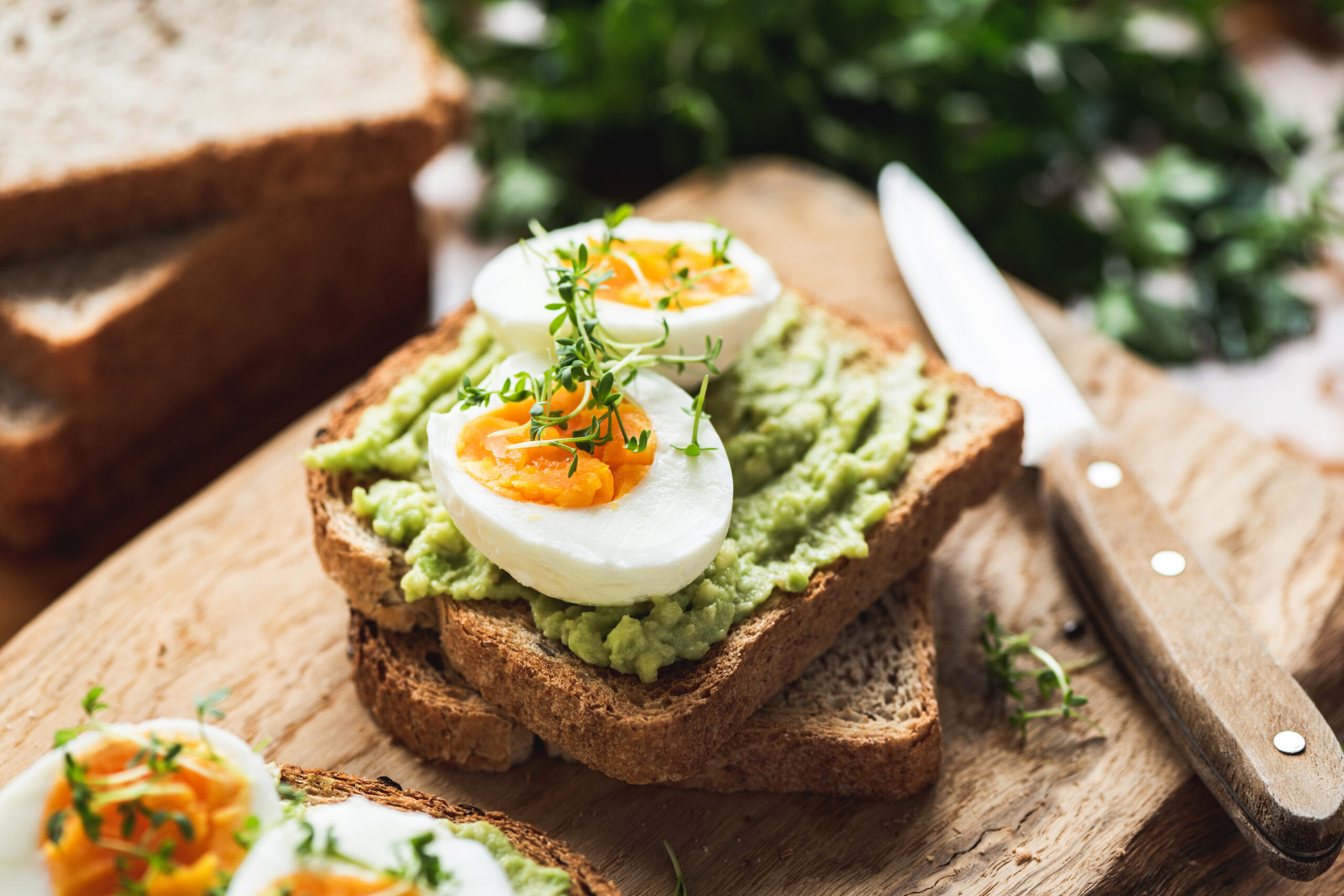 Slices of toast topped with mashed avocado, halved boiled eggs, and a sprinkle of fresh herbs. A knife rests beside the toast on a wooden cutting board, with more slices of bread and leafy greens blurred in the background.