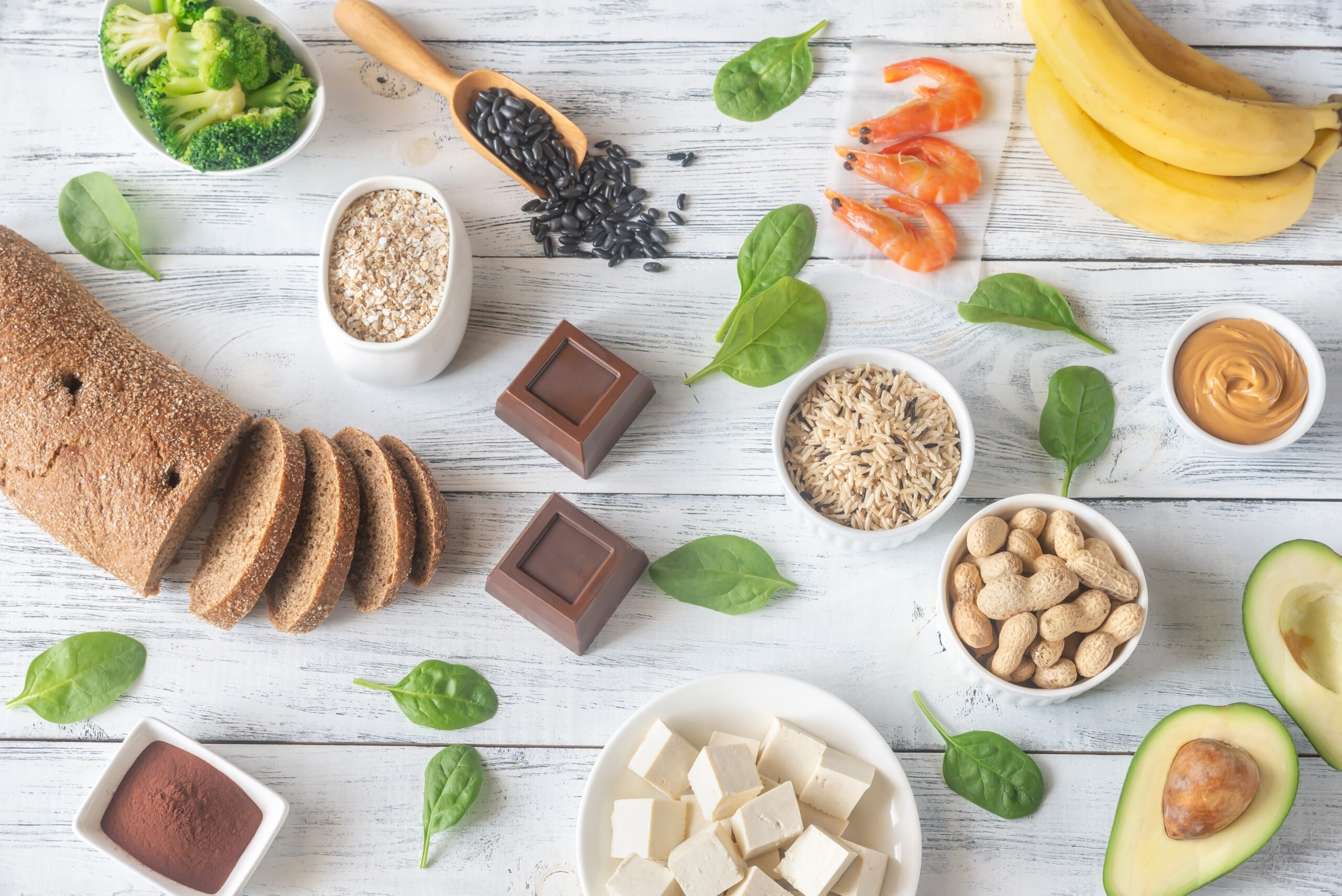 An assortment of magnesium-rich foods displayed on a wooden surface, including bread, broccoli, oats, black beans, shrimp, bananas, avocado, peanut butter, peanuts, tofu, dark chocolate, cocoa powder, spinach leaves, and brown rice.
