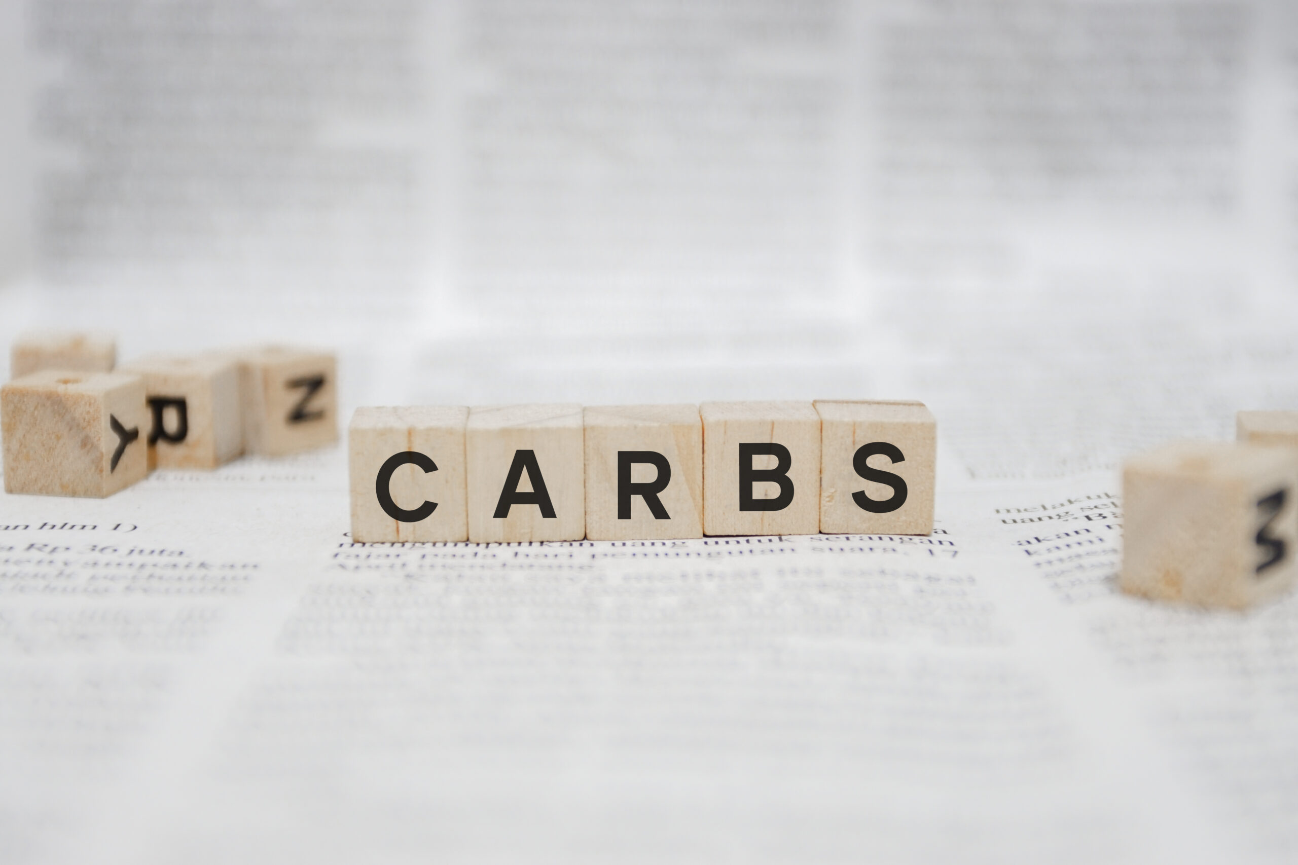 Scrabble tiles on a white surface spell out the word "CARBS." Additional scrabble tiles with letters are scattered around in the background. The surface below the tiles appears to be covered with text from a book or newspaper.