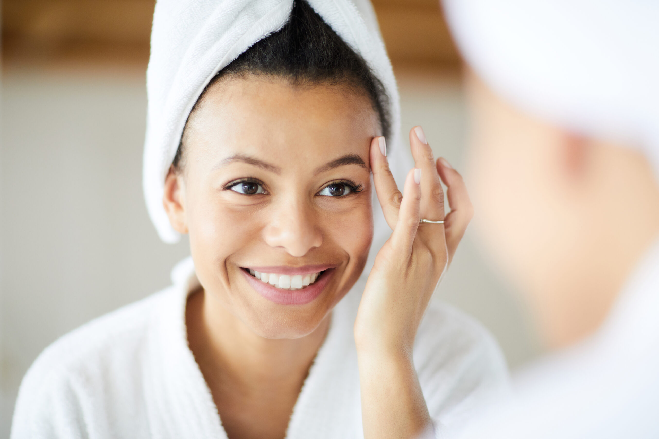 A smiling woman with a towel wrapped around her head stands in front of a mirror, touching her face gently. She is wearing a white bathrobe and appears to be in a bathroom. The background is blurred, highlighting her relaxed and happy expression.