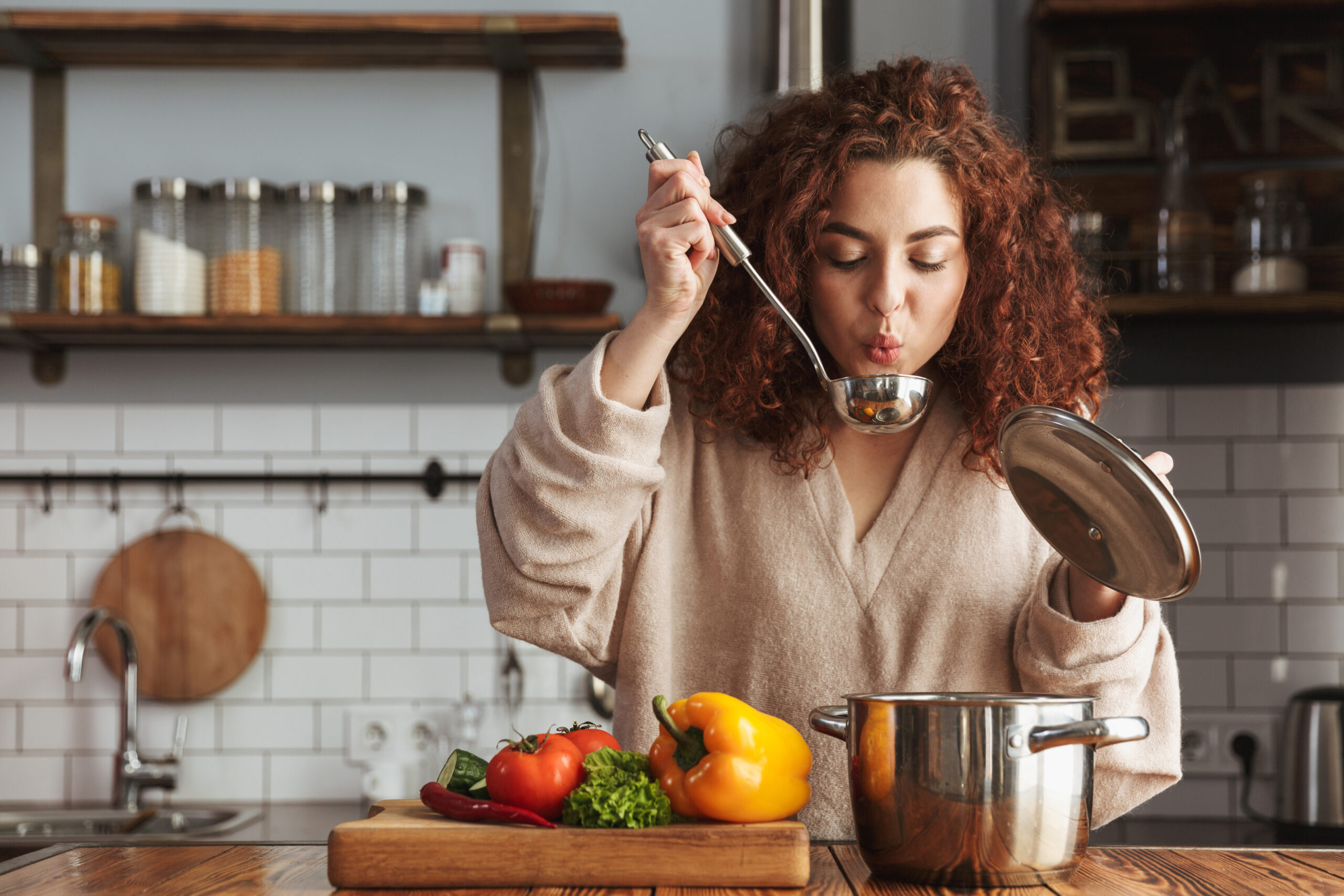 A woman with curly red hair wearing a beige sweater is blowing on a ladle filled with soup over a pot in a kitchen. She is beside a wooden cutting board with various vegetables, including bell peppers and tomatoes, and shelves with jars in the background.
