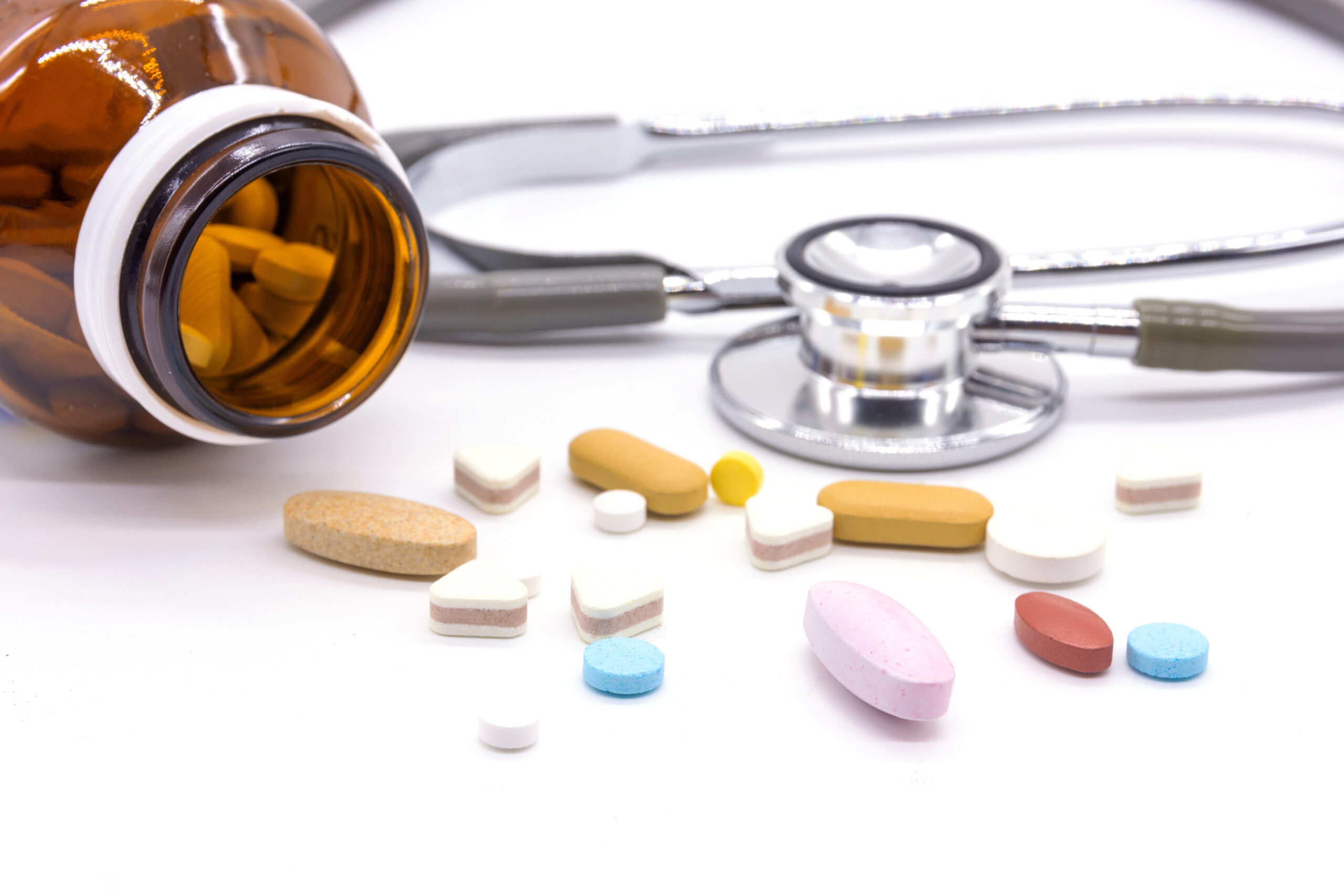 A brown pill bottle is tipped over with variously colored and shaped pills scattered out. A stethoscope lies in the background, partially in focus, suggesting a medical or healthcare theme. The white background provides a clean, clinical look.