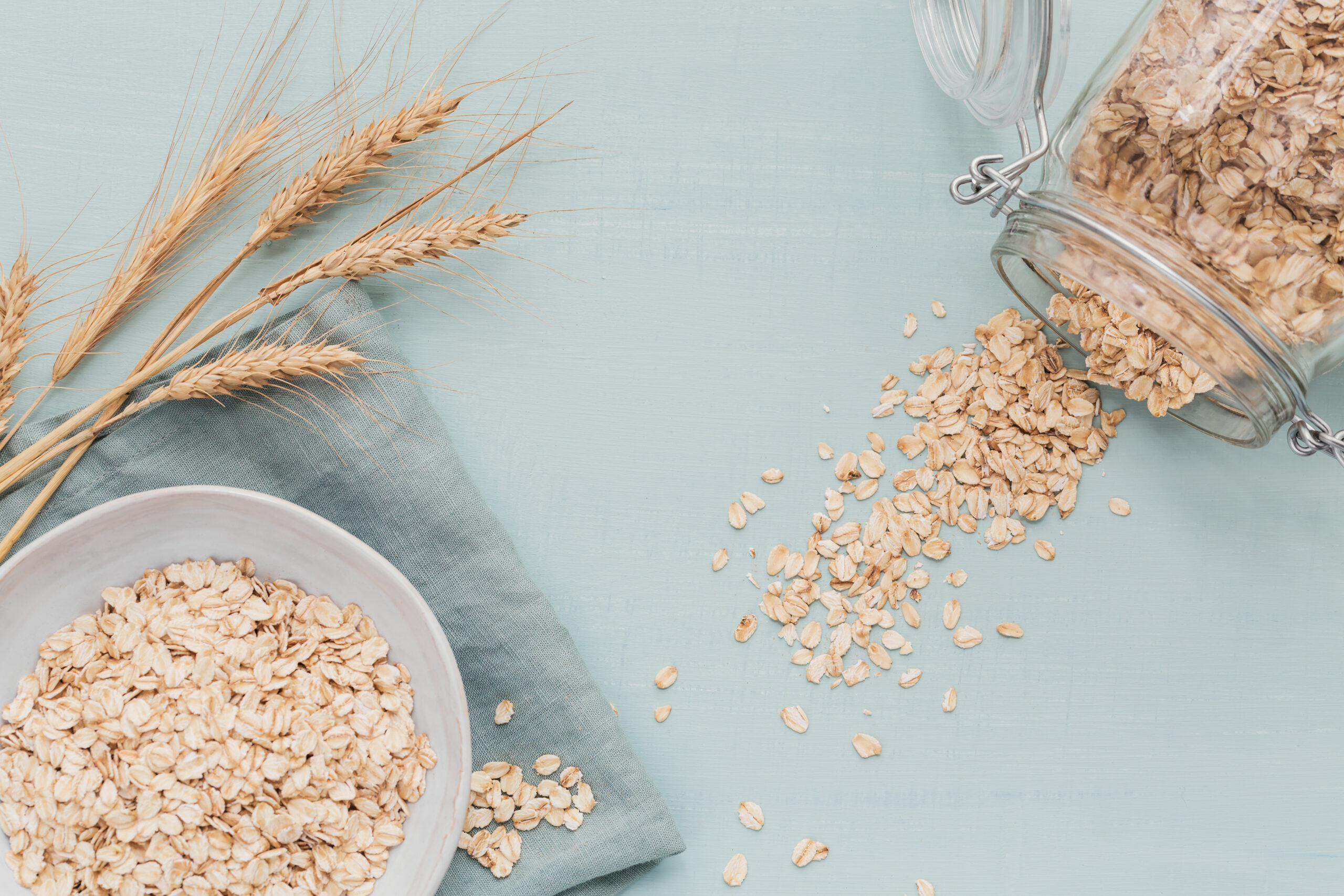A bowl of oatmeal sits on a light blue surface next to a glass jar with a metal clasp, spilling oats. Stalks of wheat are arranged on the left. A light green cloth is partially visible under the bowl and jar.