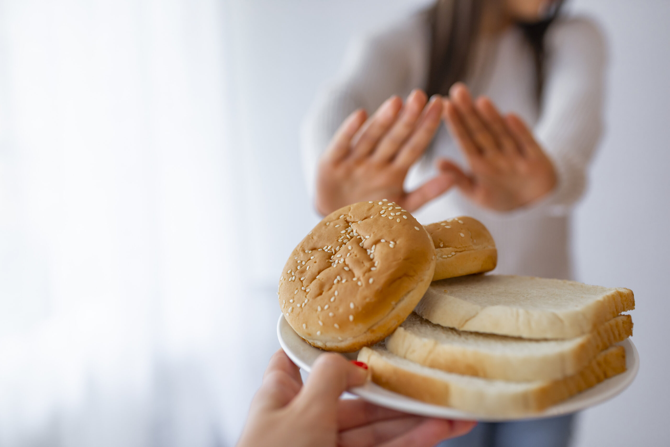 A person holds a plate with hamburger buns and slices of white bread in the foreground, offering it to a blurred figure in the background. The figure in the background has their hands raised, signaling refusal or rejection of the bread.