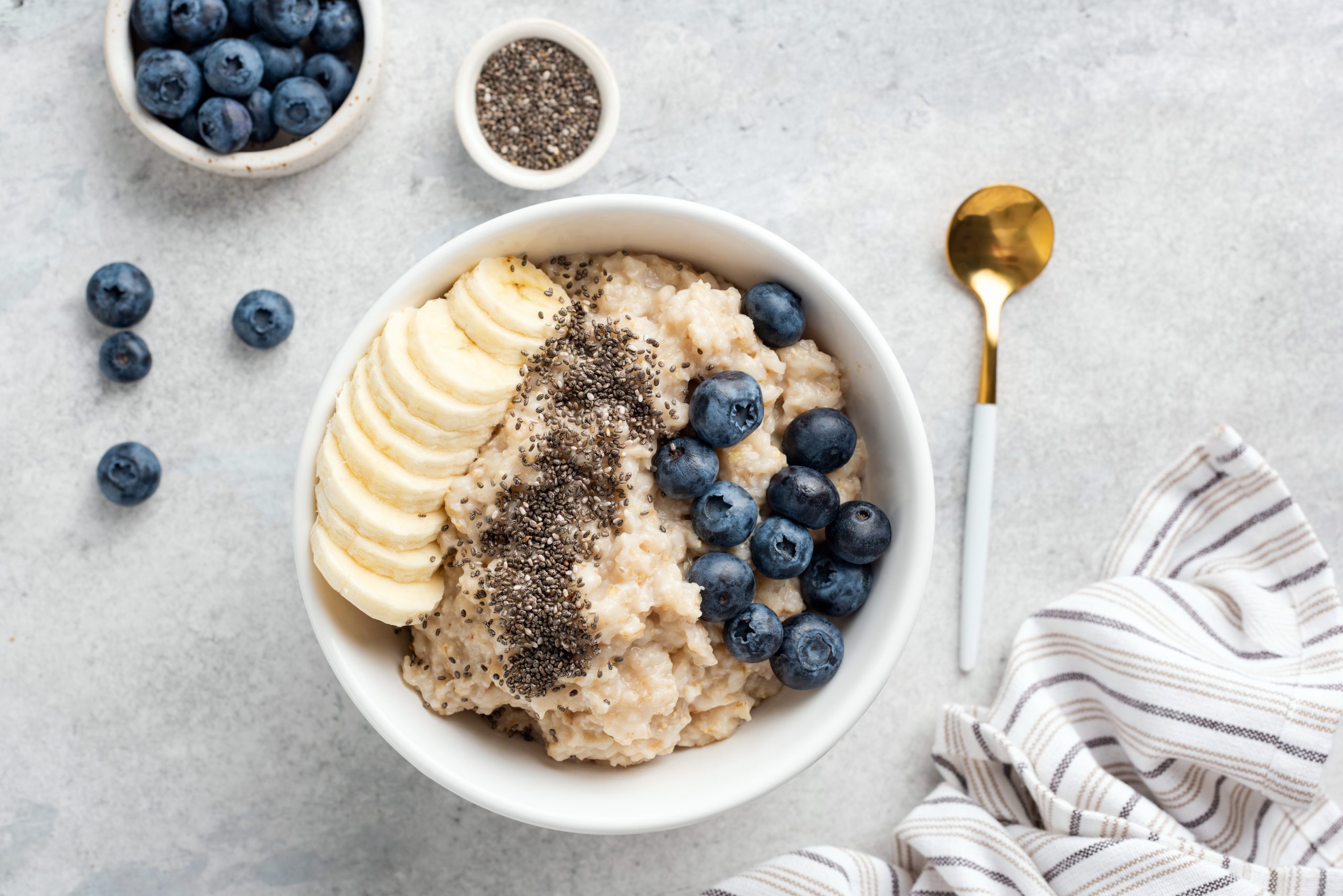 A bowl of oatmeal topped with sliced banana, blueberries, and chia seeds is on a light gray surface. Nearby, there are small bowls of blueberries and chia seeds, scattered blueberries, a gold and white spoon, and a striped cloth napkin.