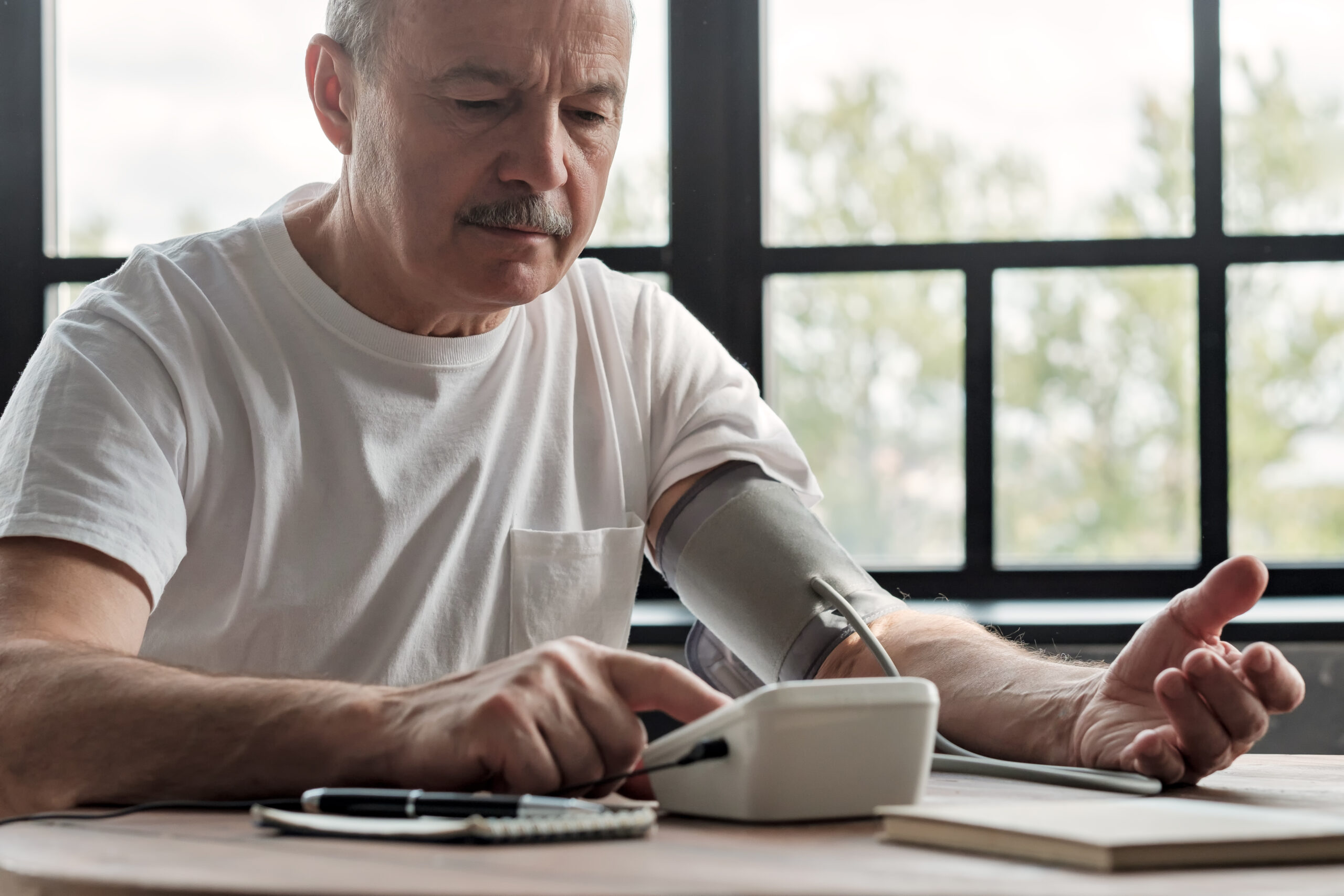 A man wearing a white t-shirt sits at a table by a large window, using a digital blood pressure monitor on his left arm. He appears focused on the device's display screen. The room is softly lit with natural light coming through the windows.