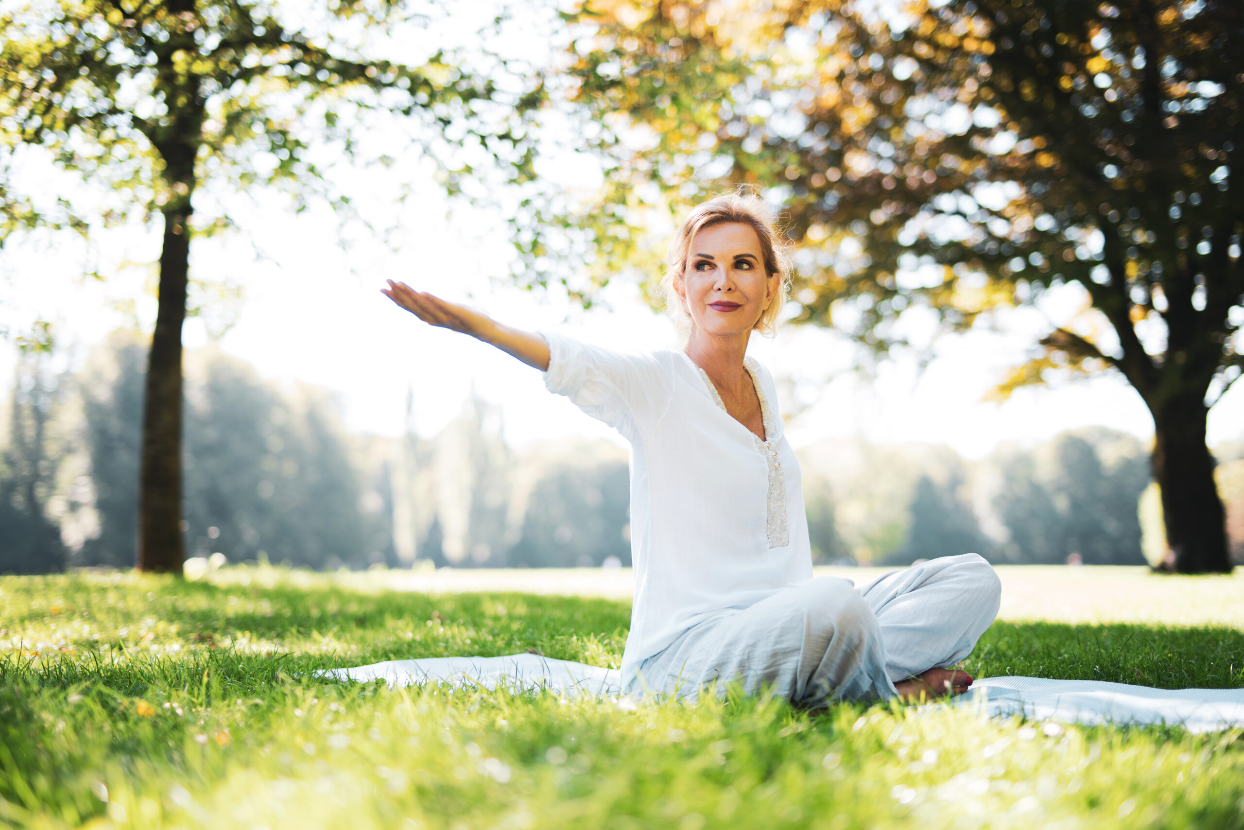 A woman sits cross-legged on a blanket in a grassy park, performing a yoga pose with one arm extended to the side. She wears a white blouse and pants, and appears calm and focused. Trees and sunlight create a serene backdrop.