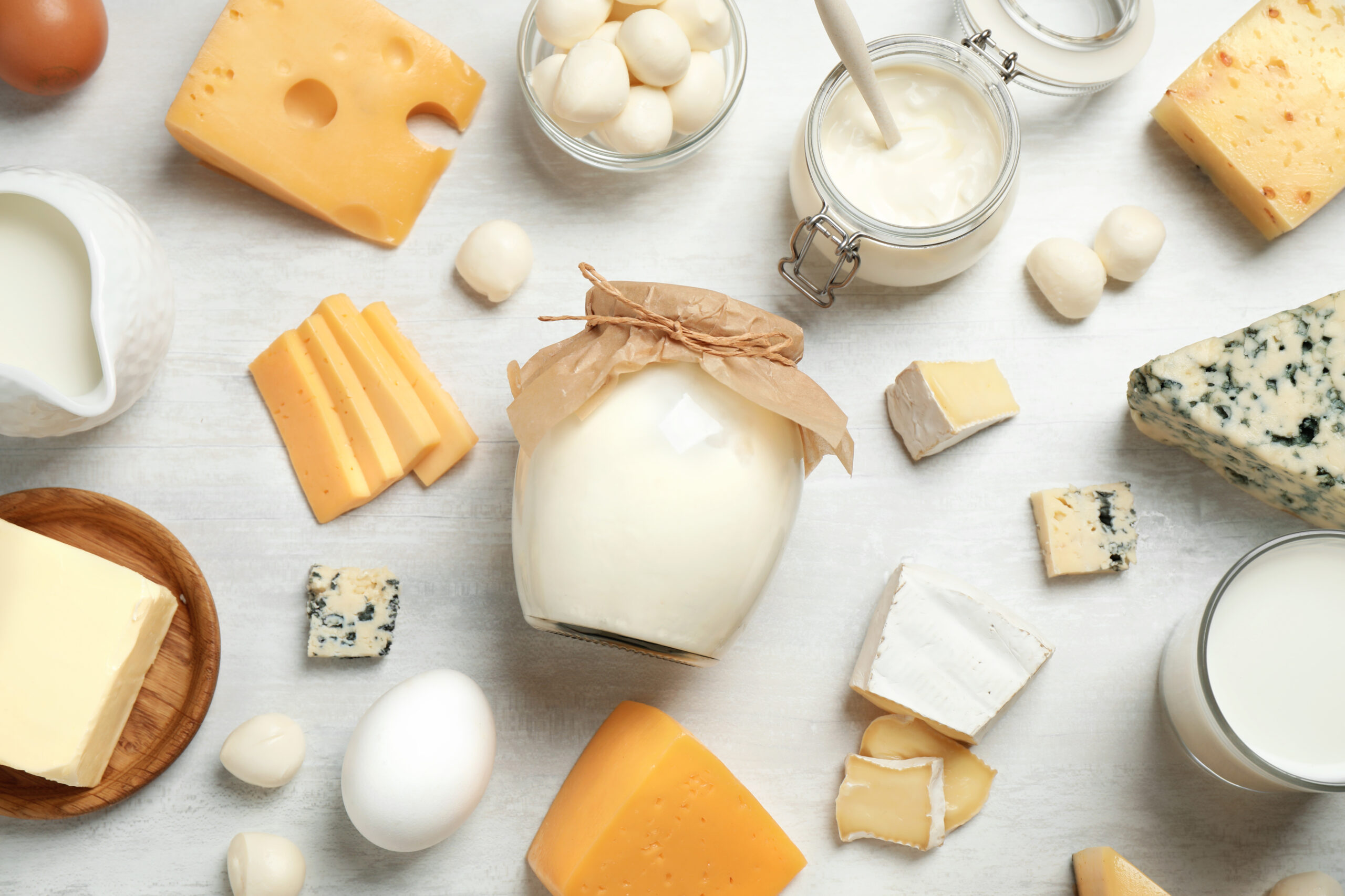A variety of dairy products arranged on a white surface. Items include different types of cheese, milk, eggs, butter in a wooden dish, and jars of cream or yogurt. The assortment showcases hard, soft, and blue cheeses, along with small round cheese balls.