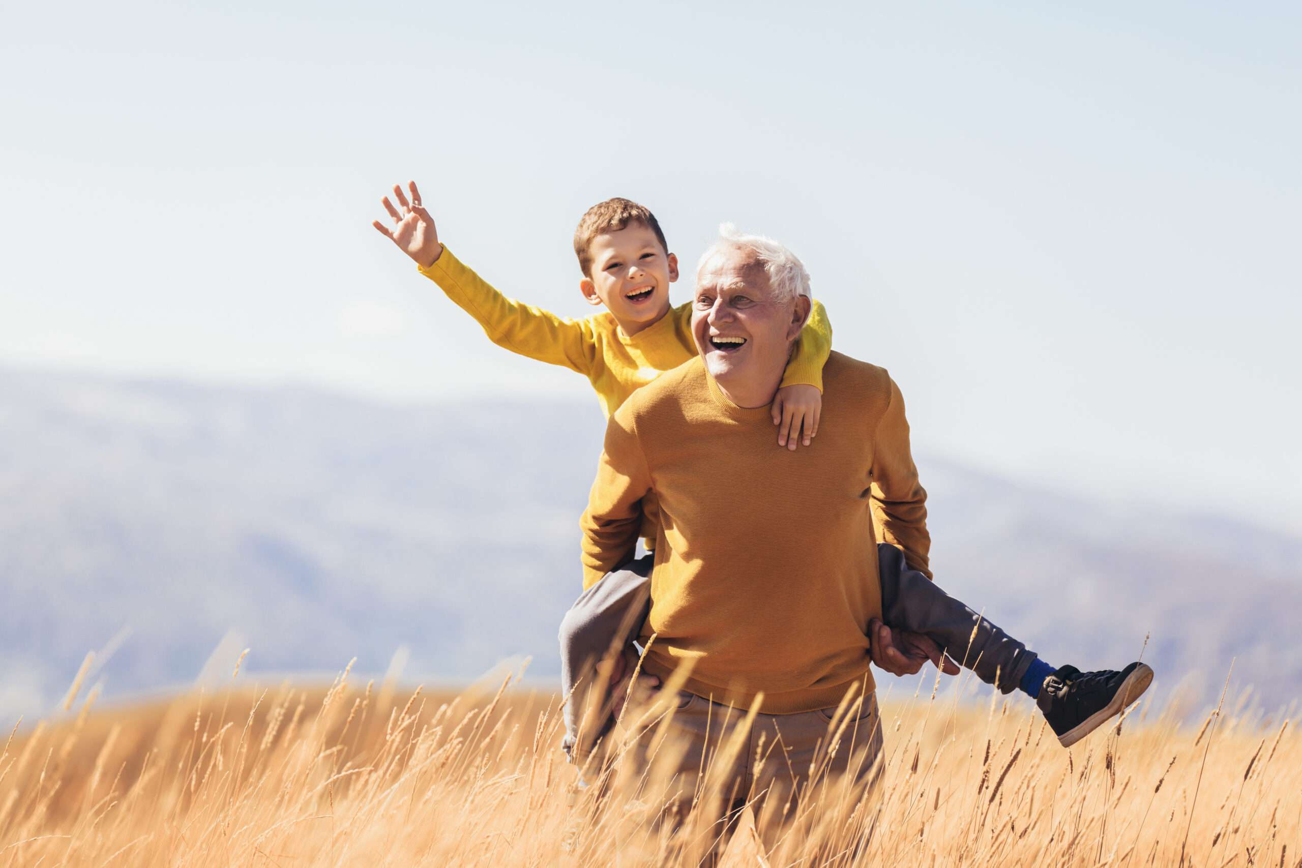 A joyful elderly man in an orange sweater gives a piggyback ride to a young boy in a matching orange sweater. They are amidst a field of tall golden grass with a clear sky and distant mountains in the background. The boy is smiling and waving his hand.