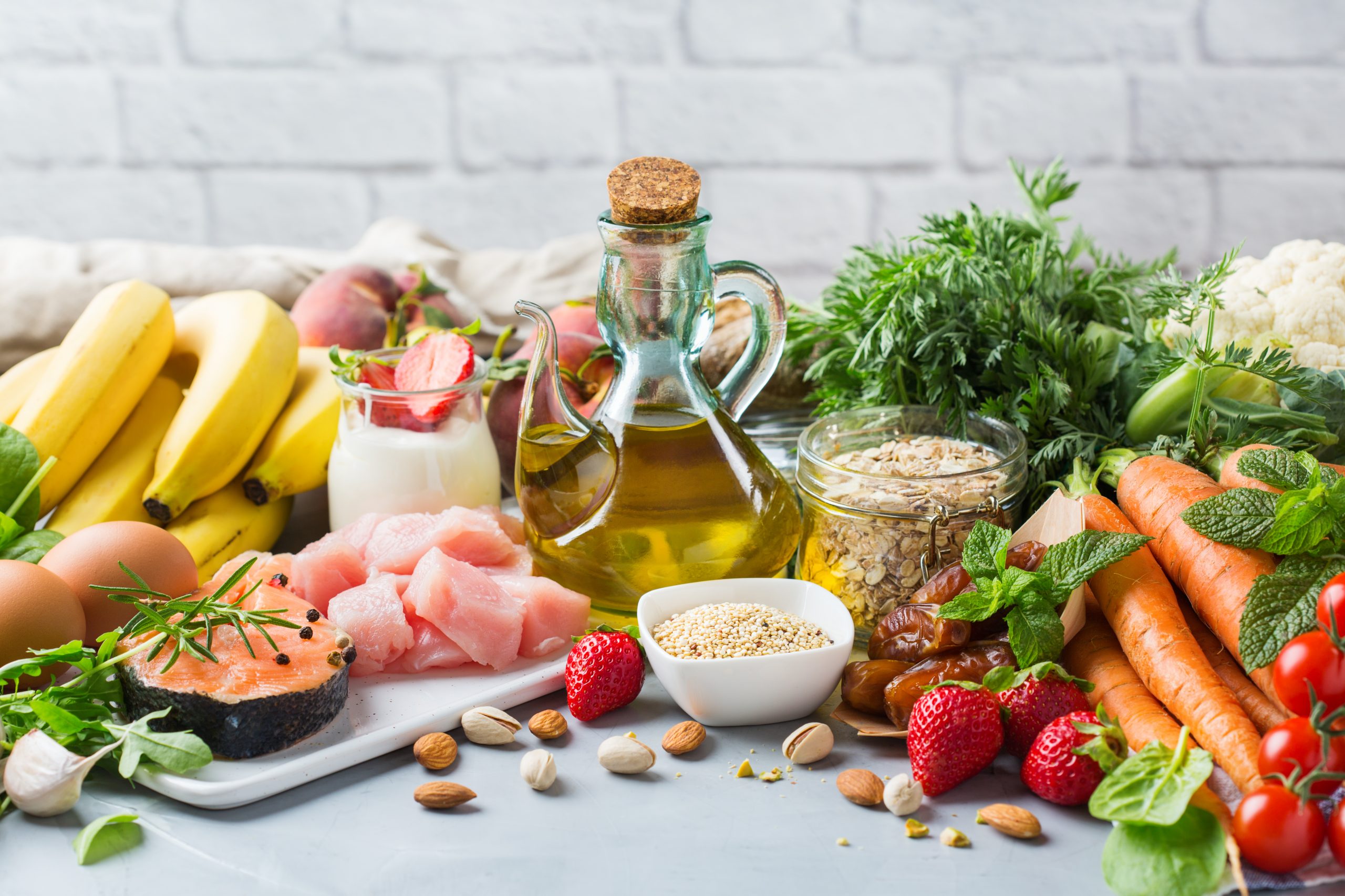 A colorful assortment of healthy foods on a table, including bananas, salmon, chicken, eggs, olive oil, yogurt with strawberries, various nuts, oats, sesame seeds, dates, strawberries, tomatoes, carrots, cauliflower, and fresh herbs against a white brick wall.