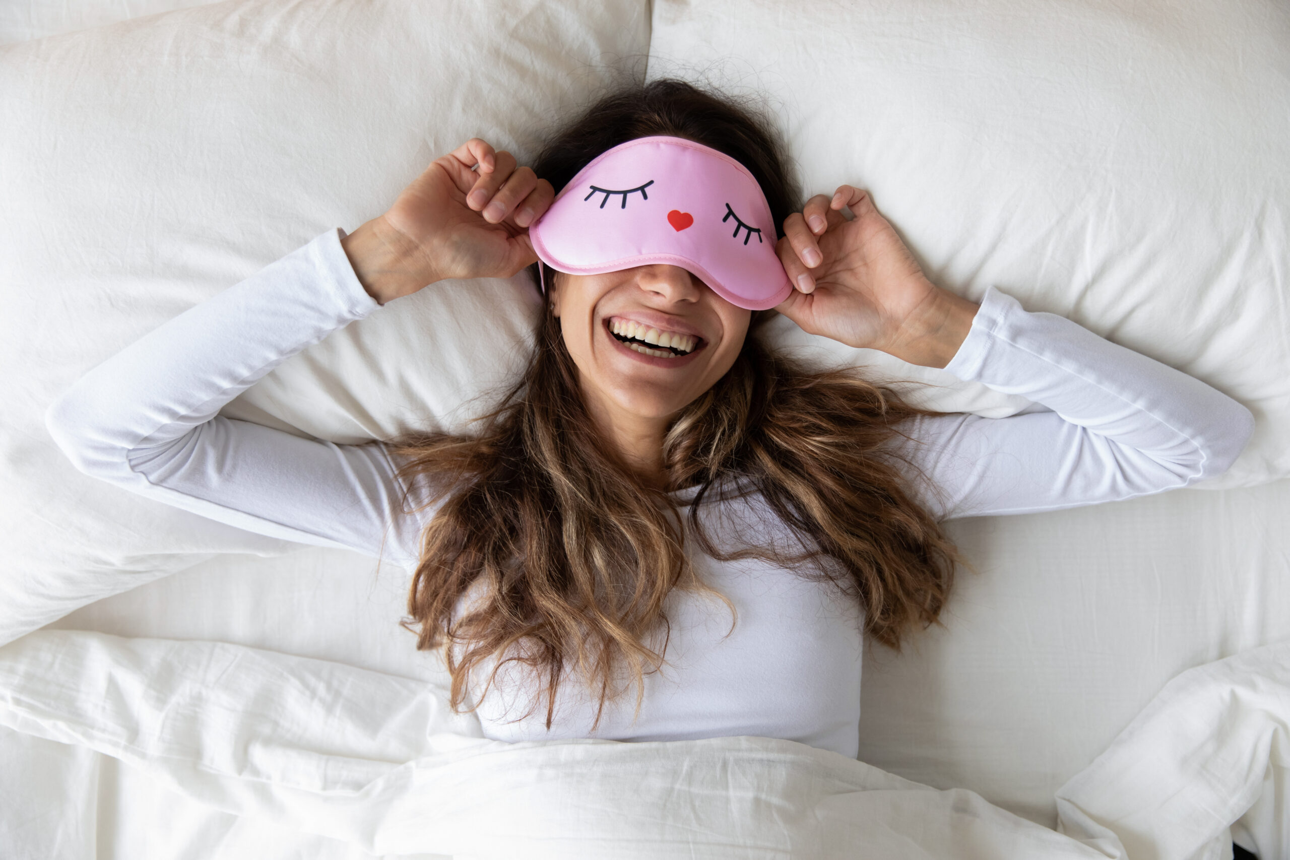 A woman is lying in bed, smiling widely while lifting a pink sleep mask decorated with cartoon eyes and a tiny heart. She is wearing a long-sleeve white shirt and has long, wavy hair spread across the pillow.