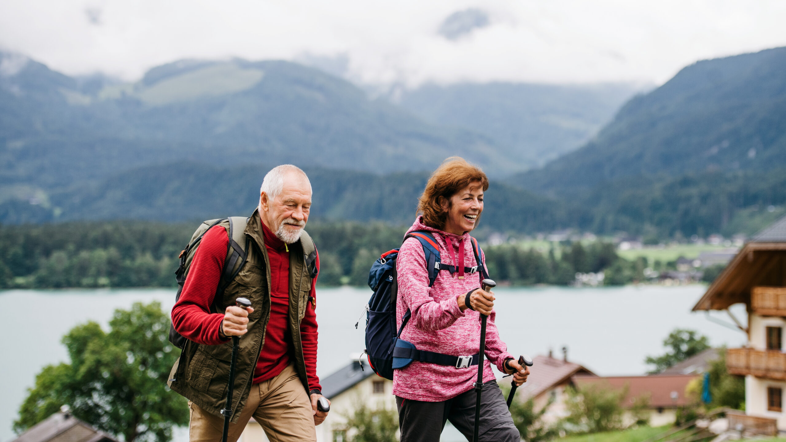 An elderly couple enjoys a hike in a mountainous region. Both are using hiking poles and wearing backpacks and outdoor clothing. The background features a scenic view of hills, a lake, and scattered buildings. The sky is cloudy, adding to the serene atmosphere.