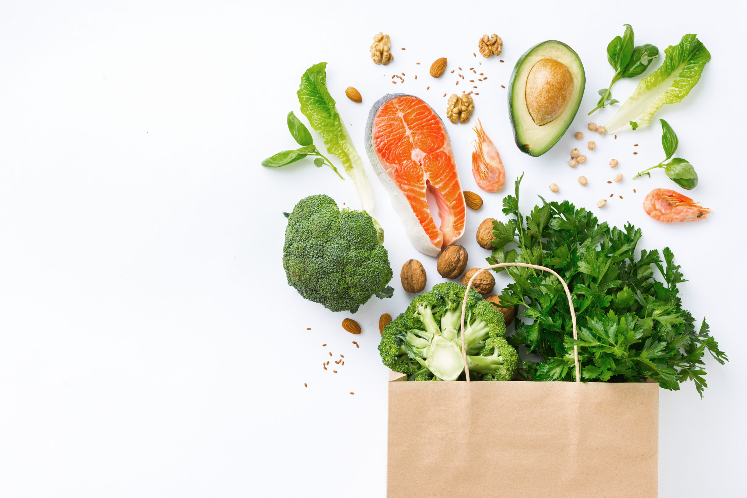 A brown paper bag on a white background, overflowing with healthy foods, including broccoli, salmon, avocado, shrimp, walnuts, lettuce, leafy greens, chickpeas, flaxseeds, and fresh herbs. The items are artfully arranged, spilling out of the bag.