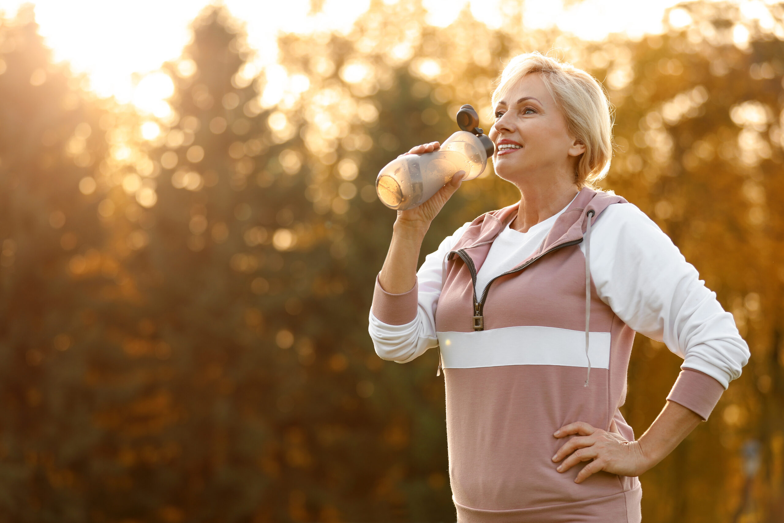 A woman with short blonde hair wearing a pink and white sporty hoodie smiles while holding a clear bottle and drinking from it outdoors. She stands against a backdrop of trees with warm sunlight filtering through, giving the scene a serene and cheerful atmosphere.