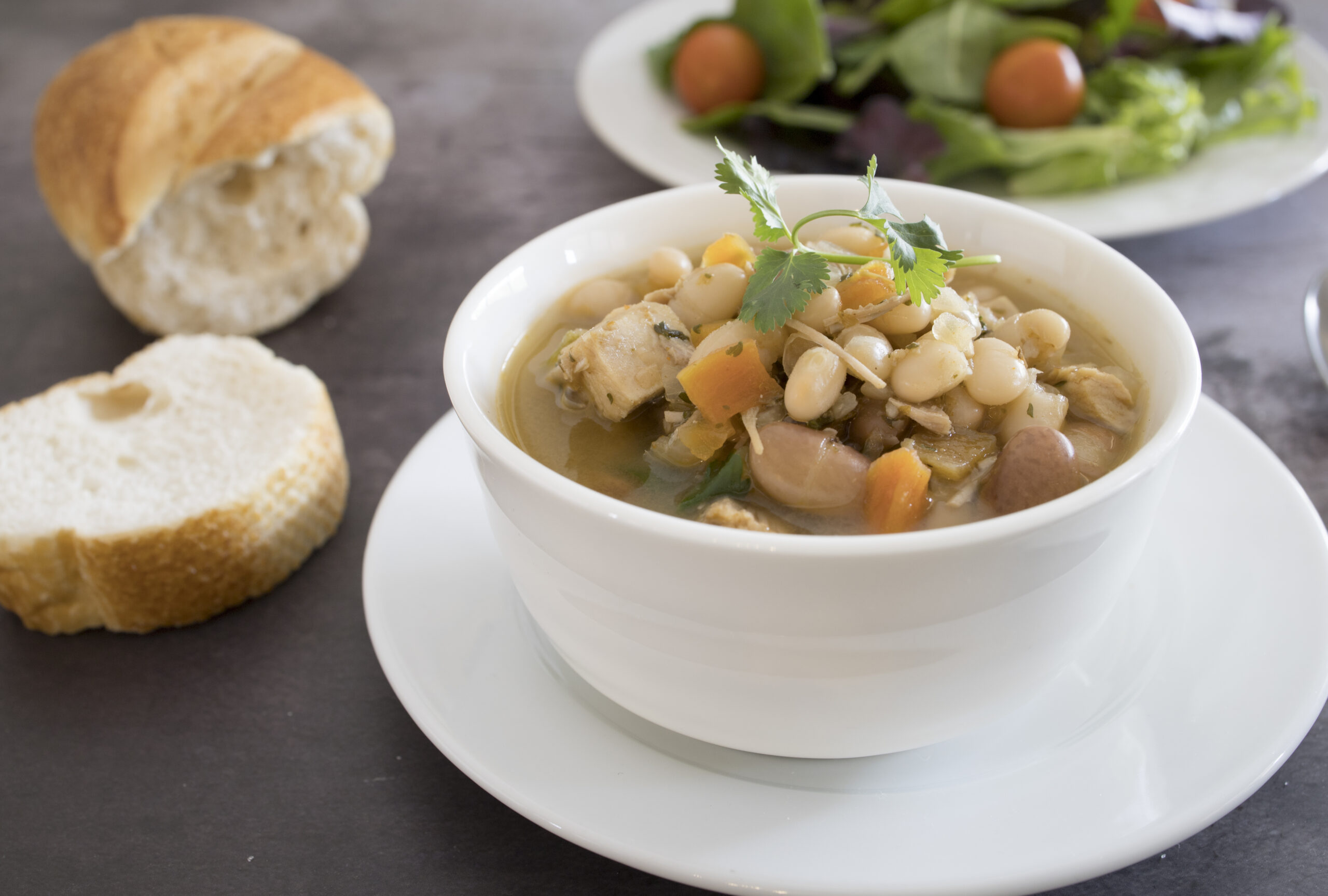 A bowl of white bean and vegetable stew garnished with cilantro, served on a white plate next to a piece of sliced bread. In the background, there is a salad with mixed greens and cherry tomatoes on a white plate. The table surface is dark gray.