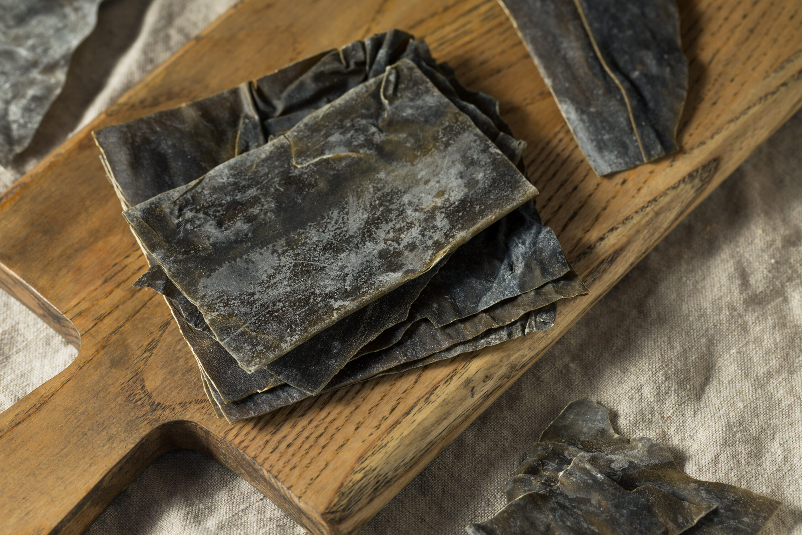 Dried kelp sheets stacked on a wooden cutting board with some pieces scattered around on a light fabric surface. The kelp appears dark and rough-textured.