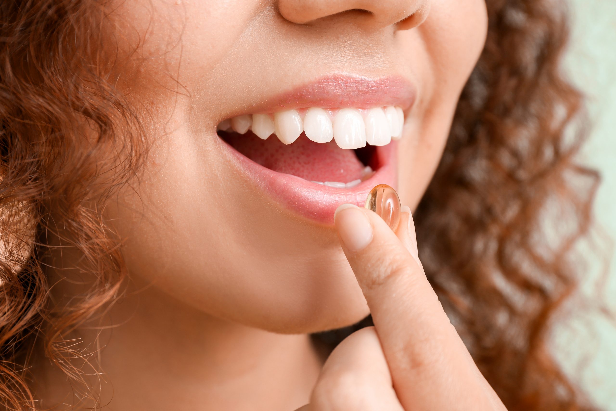 A close-up of a person with curly hair holding a gel capsule pill near their mouth. The person has a smile showing white teeth, and their lips are slightly parted, ready to take the pill. The background is blurred with a light greenish hue.