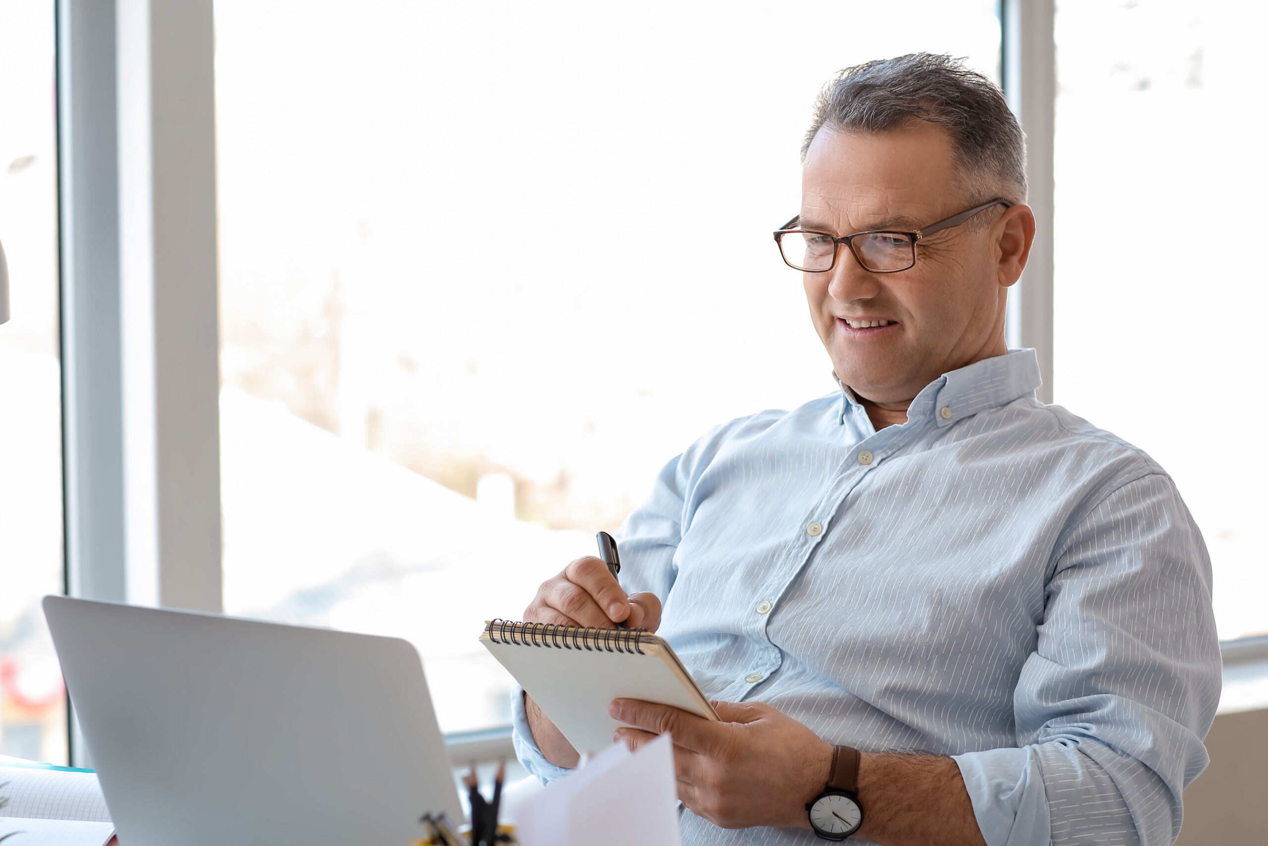A man with short, graying hair and wearing glasses is sitting at a desk in front of a laptop. He is smiling while taking notes in a spiral notebook. He is dressed in a light blue, button-up shirt and wearing a wristwatch. The background shows a bright window.