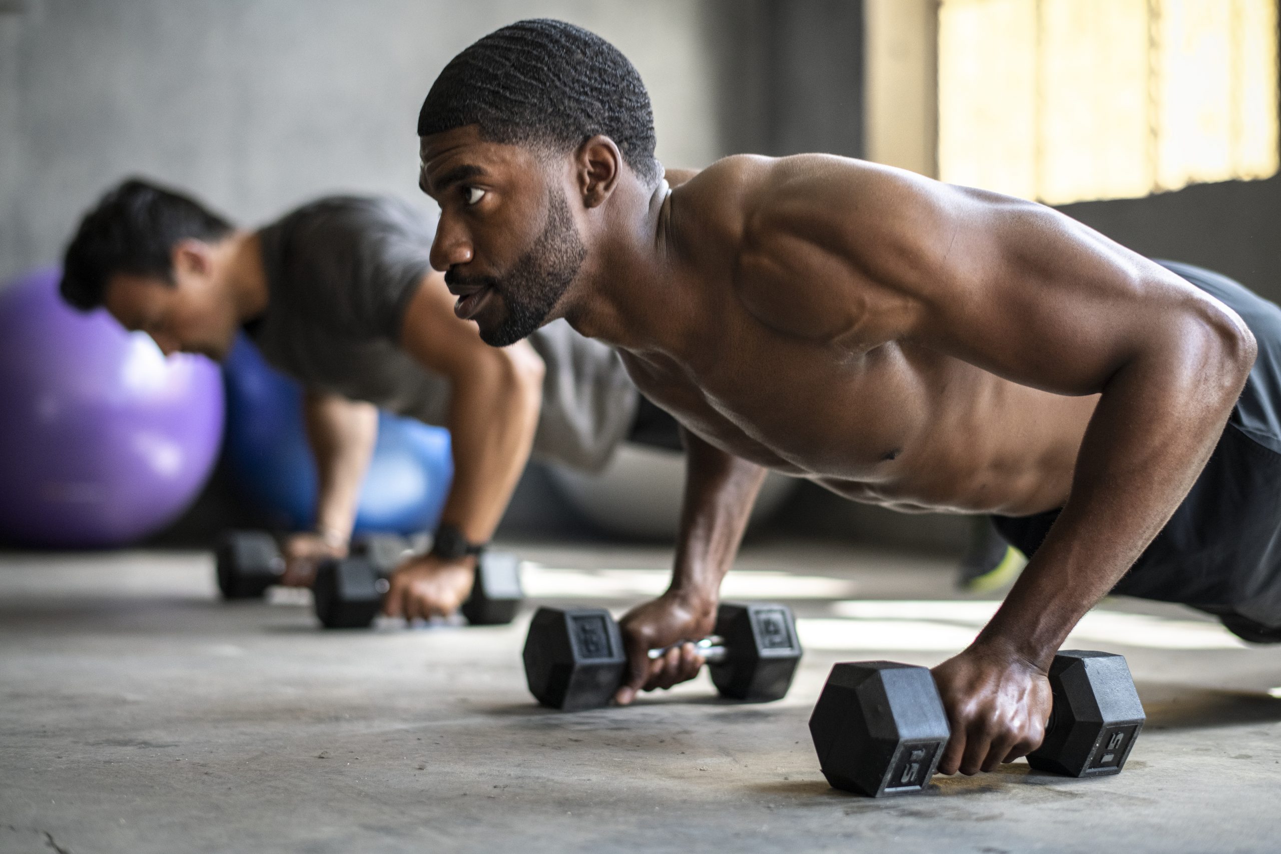 Two men are performing push-ups while gripping dumbbells in a gym. The man in the foreground is shirtless and has a focused expression. Both appear to be engaged in an intense workout. The background includes an exercise ball and a slightly blurred second man.