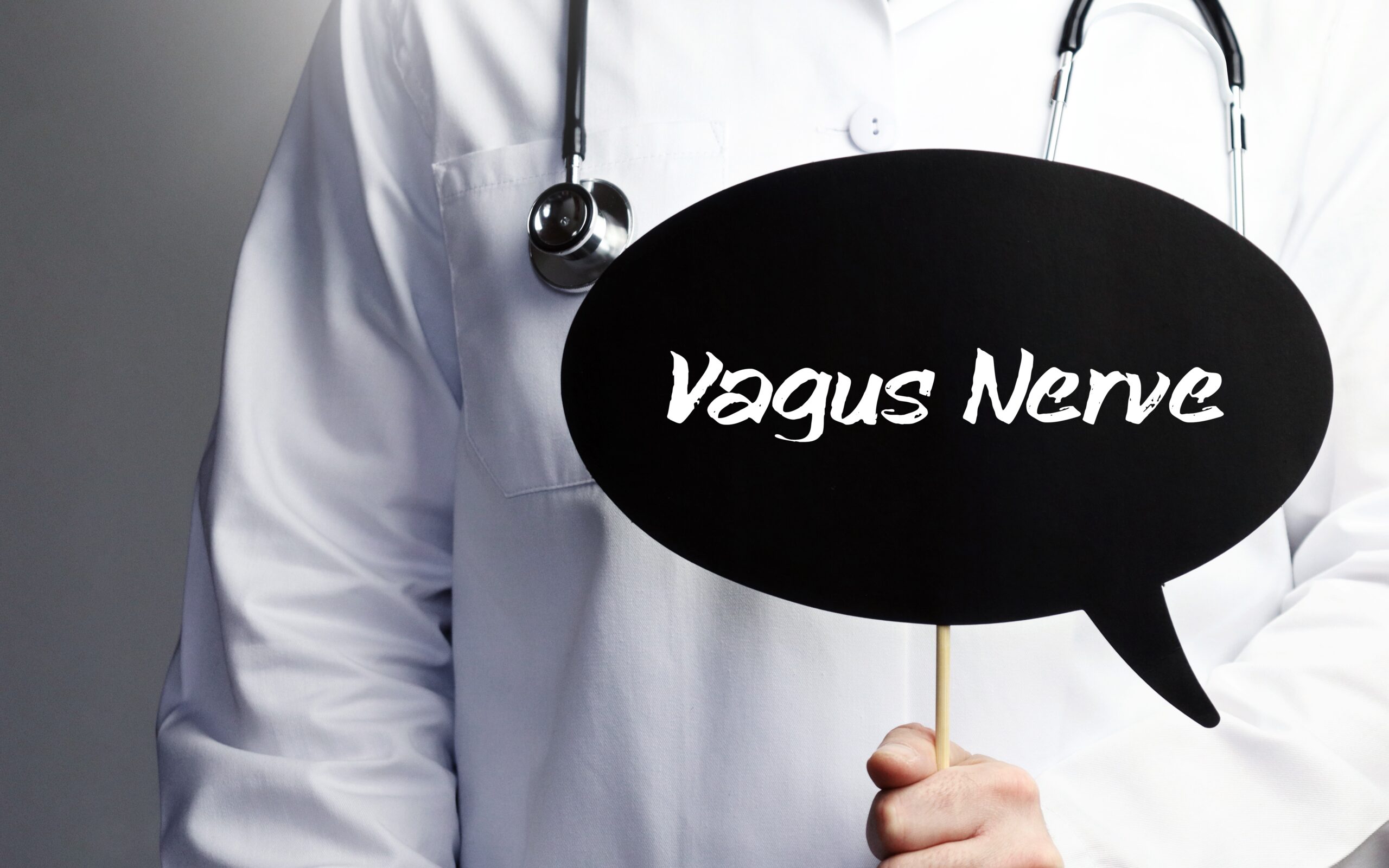 A person in a white lab coat, with a stethoscope draped around their neck, holds a black speech bubble sign with the words "Vagus Nerve" written on it in white text. The background is plain and gray.