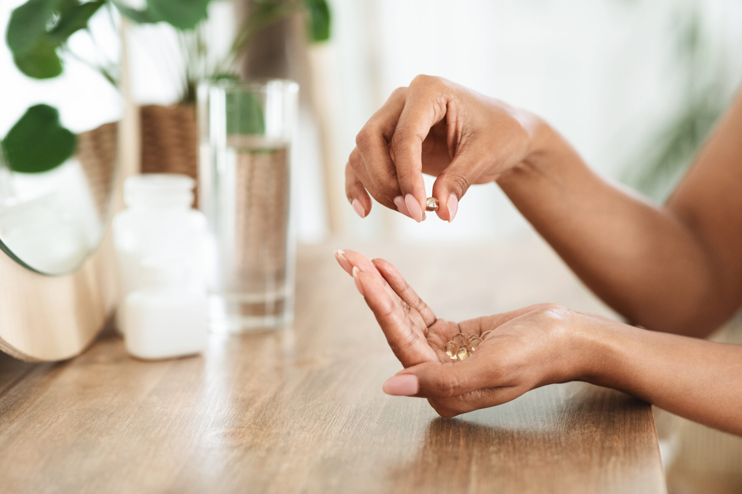 Close-up of a person holding several vitamin capsules in one hand and picking one up with the other hand, near a wooden table. The background includes a glass of water, a bottle, and a plant.