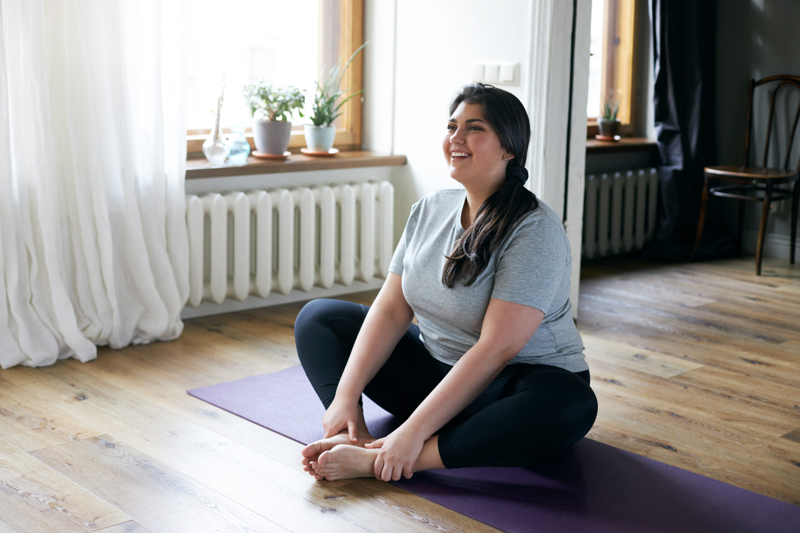 A woman sits on a purple yoga mat in a butterfly stretch position, smiling and looking relaxed. She is wearing a gray T-shirt and black leggings. There are potted plants on a windowsill behind her, and soft natural light fills the room.