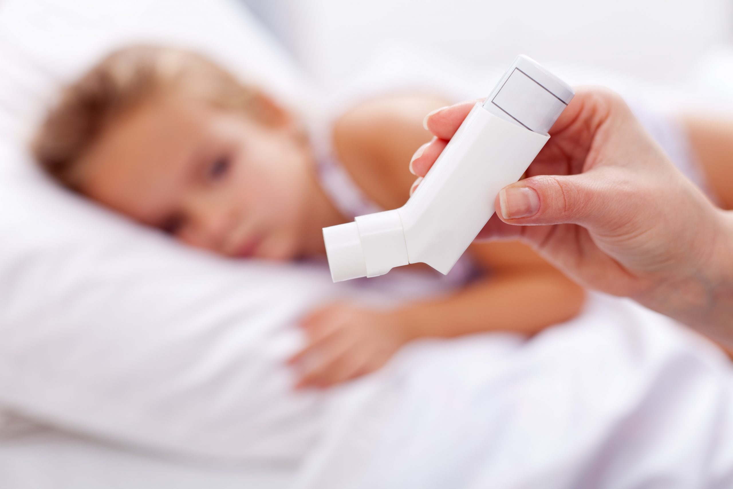 A close-up of a hand holding an inhaler in the foreground. In the blurred background, a child is lying on a bed, appearing tired or unwell, with their head resting on a pillow—highlighting how Omega-3 fatty acids can reduce asthma symptoms in kids.