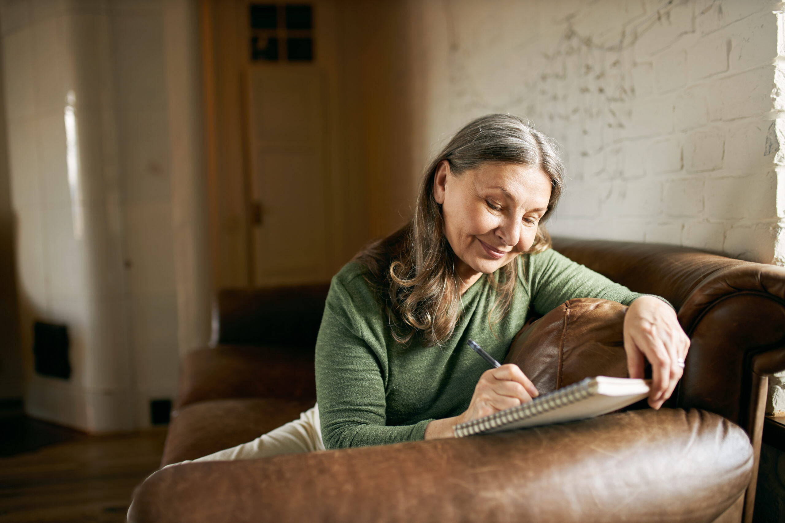 A woman with long gray hair sits comfortably on a brown leather sofa, writing in a notebook with a content expression. She is wearing a green sweater, and the setting appears to be a cozy, warmly lit room with white brick walls.