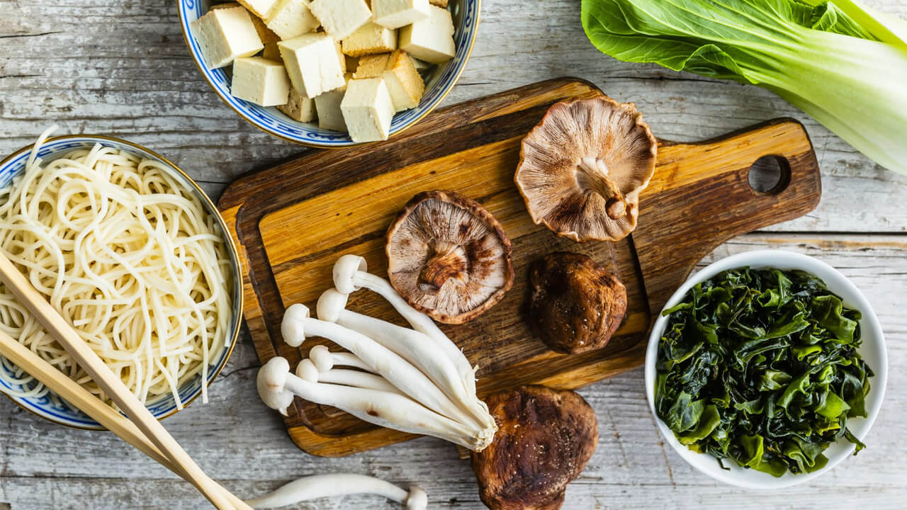 A wooden cutting board with assorted mushrooms, tofu cubes, and bok choy next to bowls of noodles, leafy greens, and enoki mushrooms on a rustic wooden table. Chopsticks rest on the bowl of noodles.